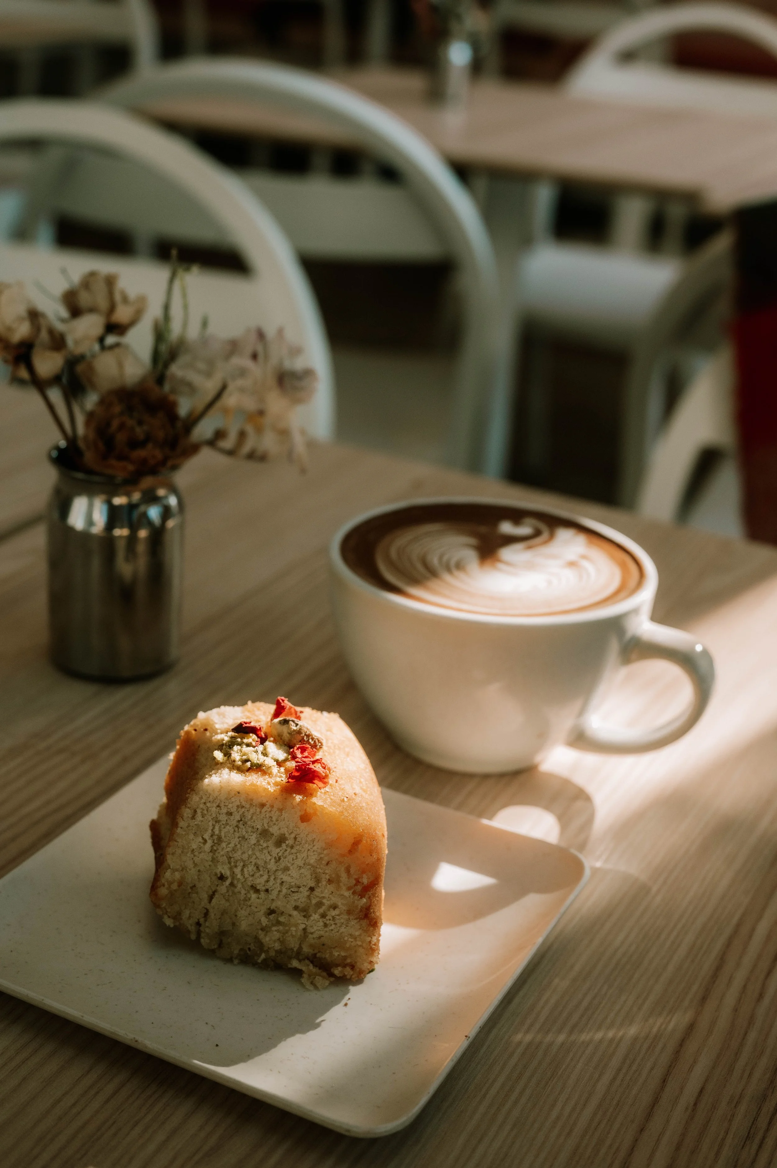 A slice of cheesecake on a white plate, with a small muffin topped with red and green garnish in the foreground. In the background, a cup of coffee with latte art and a small metal vase with dried flowers on a wooden table.