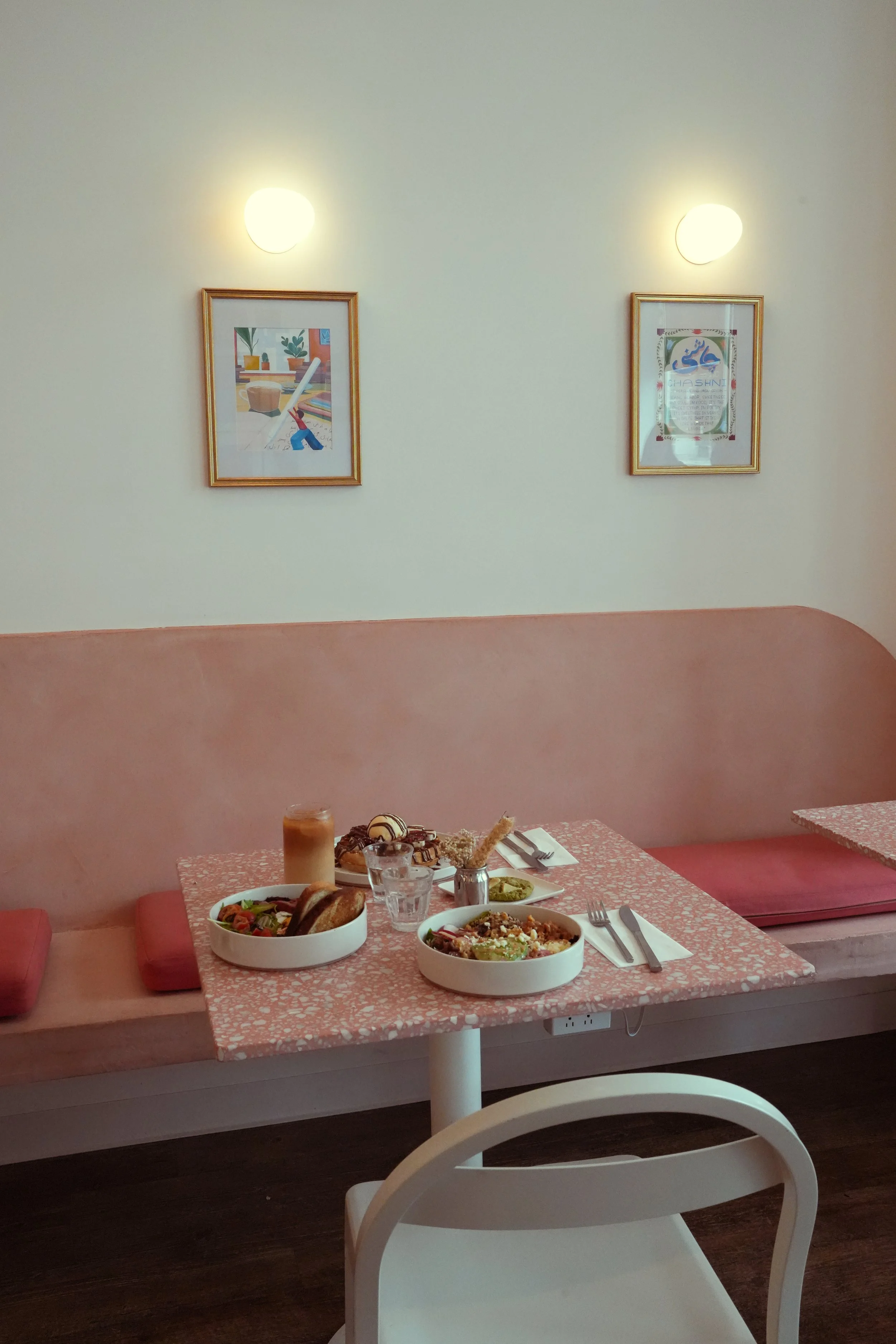 A pink-speckled table set with bowls of salad, bread, and other dishes, with a drink on the side, in a cozy restaurant with framed artwork on cream-colored walls and soft lighting.