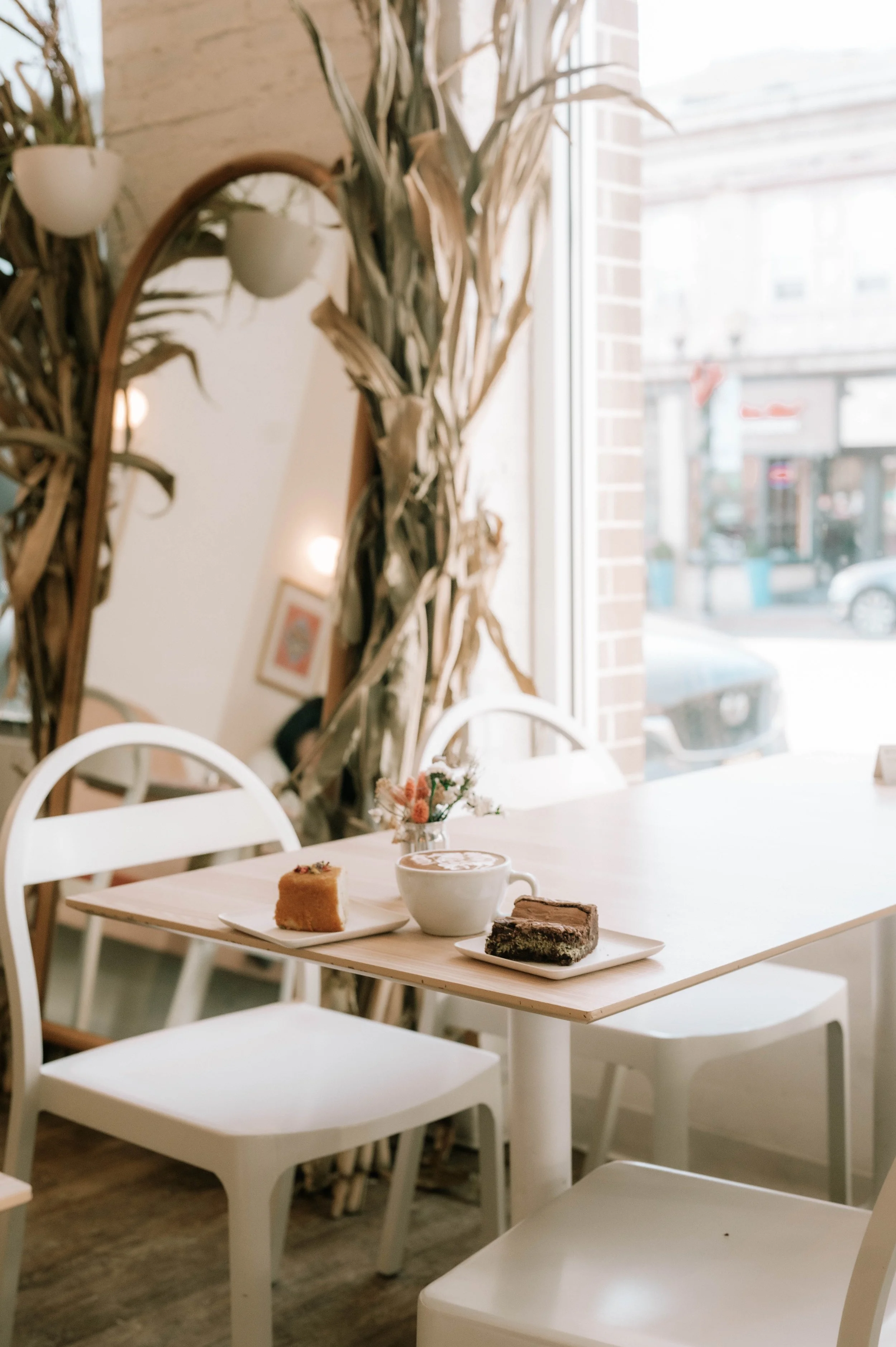 White table with three plates of desserts and a cup of coffee in a cozy cafe with white chairs and large window bringing in natural light.