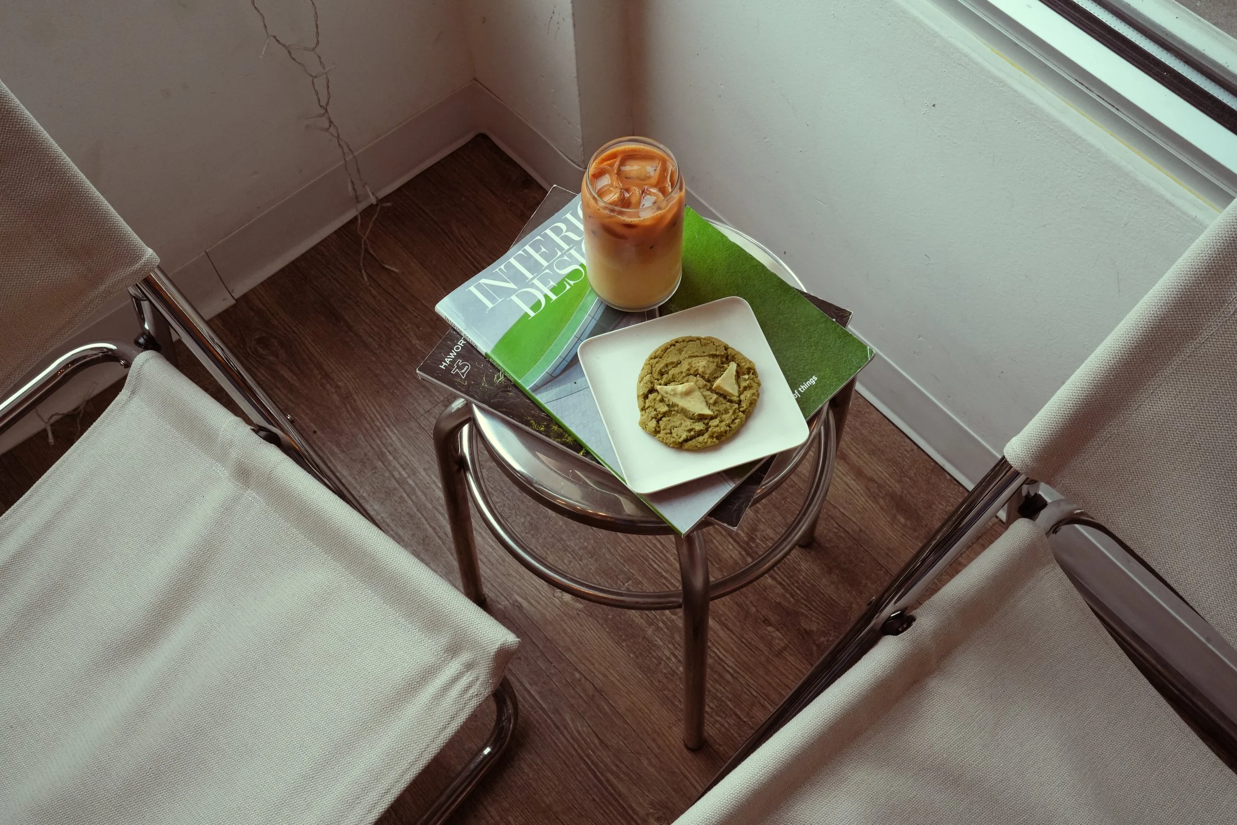 A small round glass of iced coffee on a table next to a white square plate with a large cookie on it. The table also holds two magazines and is surrounded by white cushioned chairs near a window.