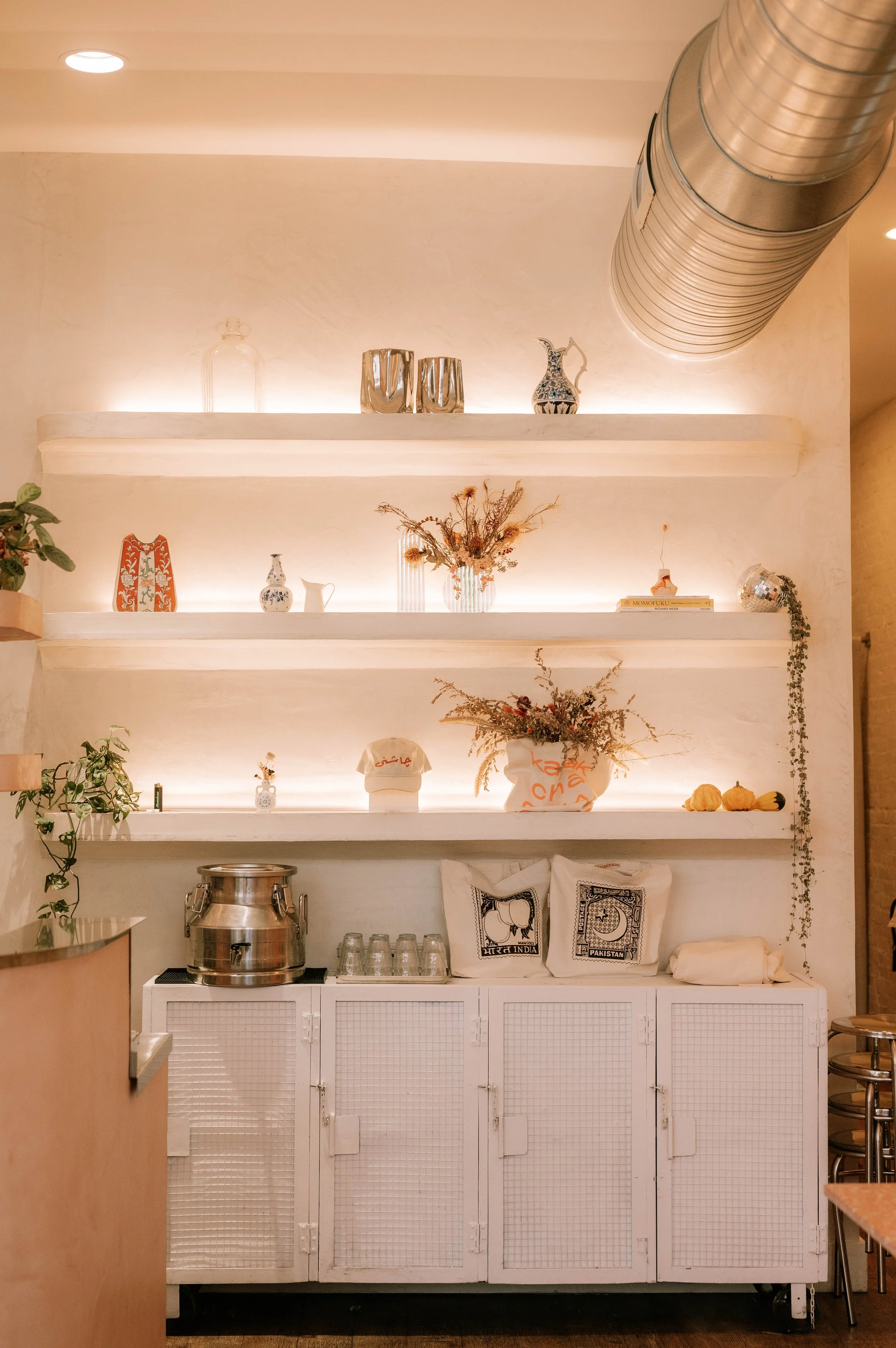 White kitchen shelves with decorative items including vases, a pitcher, books, dried flowers, and a disco ball, with a ventilation duct overhead and a counter with a silver container and glasses.