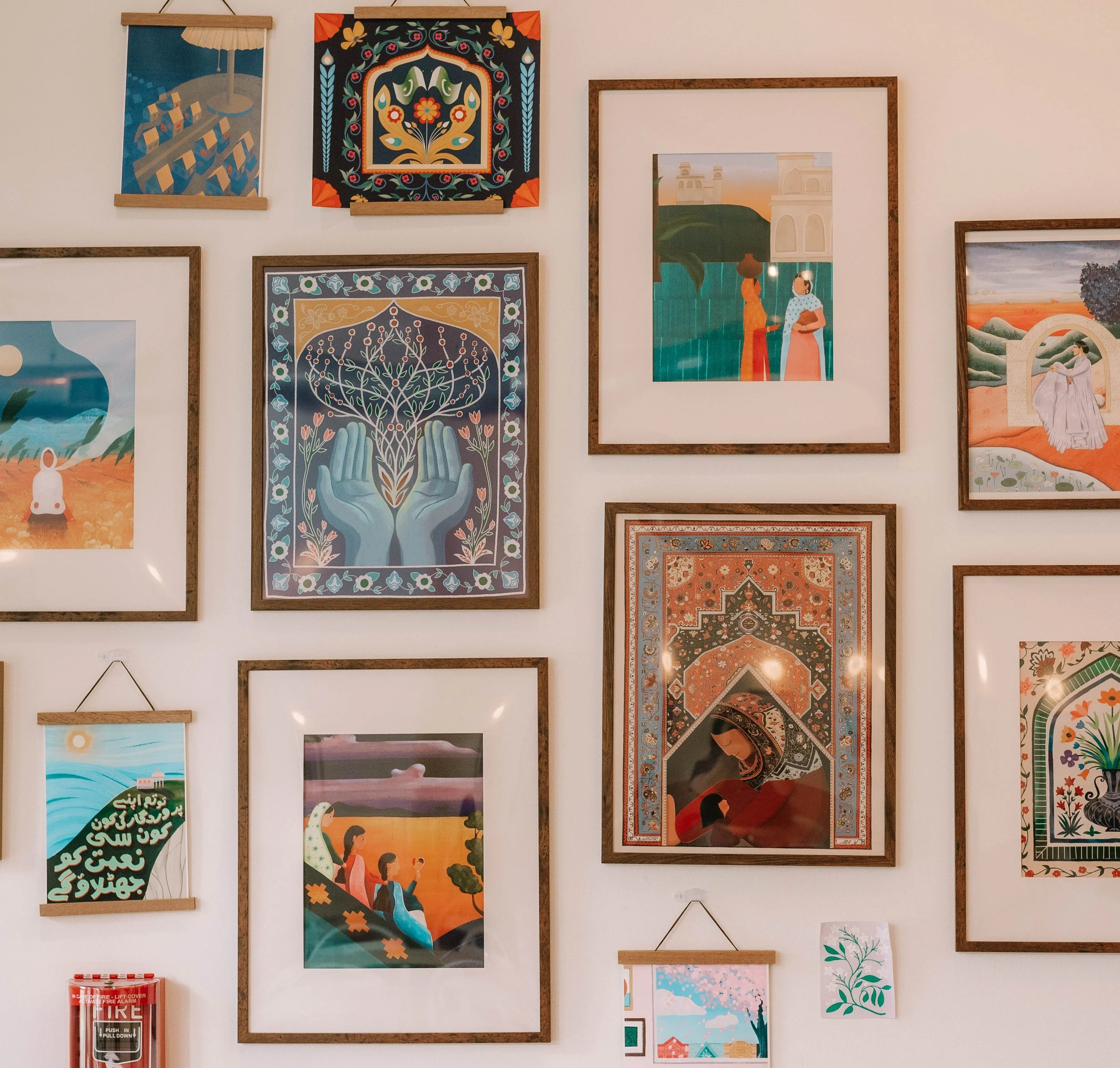 Collection of framed artwork and decorative pieces on a white wall, including illustrations of landscapes, people, and abstract patterns, with a fire alarm box at the bottom left.