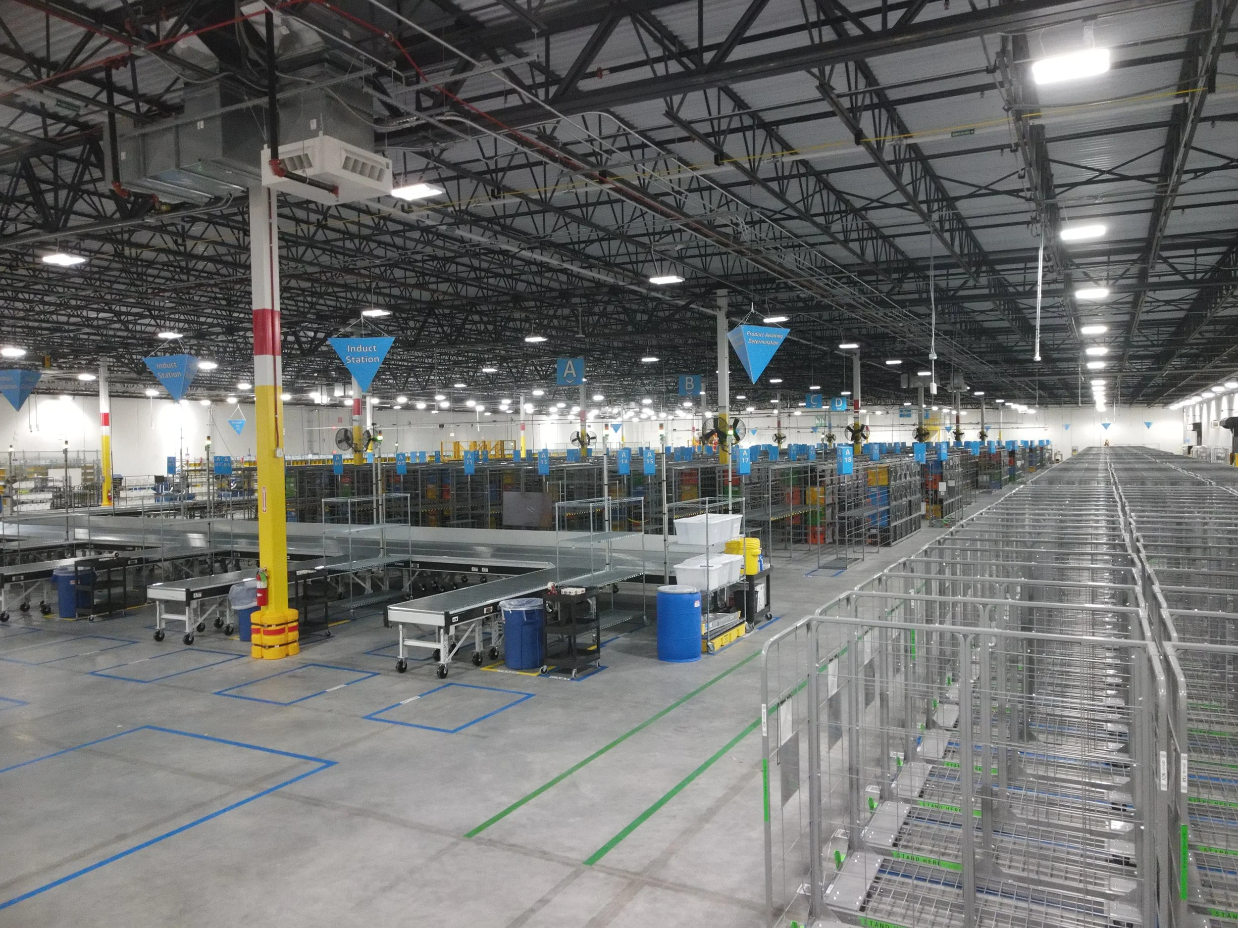 Empty warehouse or store with high ceiling, fluorescent lighting, metal shelving, shopping carts, and signs indicating handles and product areas.