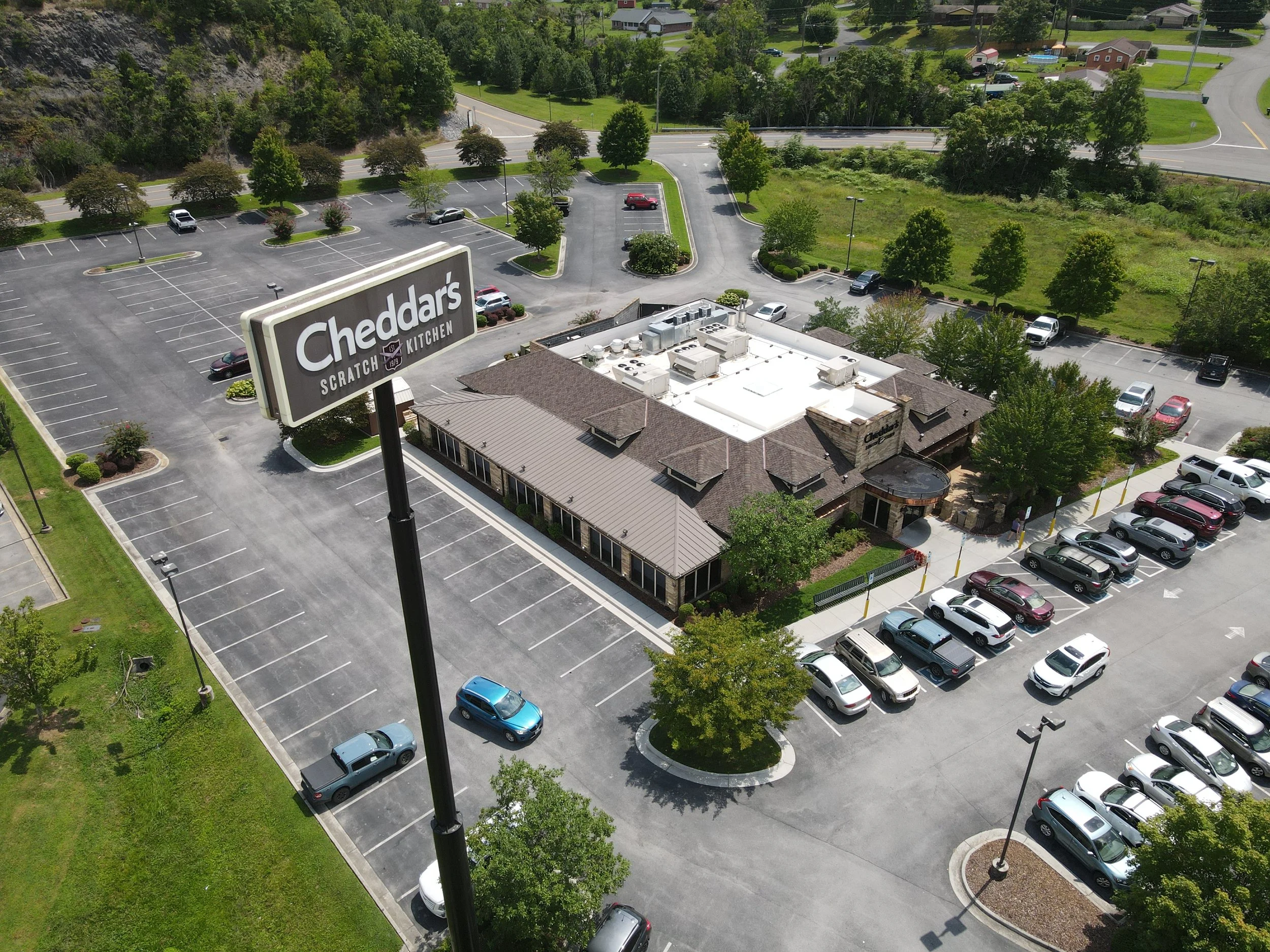 Aerial view of a parking lot outside Cheddar's Scratch Kitchen restaurant with several parked cars, surrounding trees, and nearby residential houses.