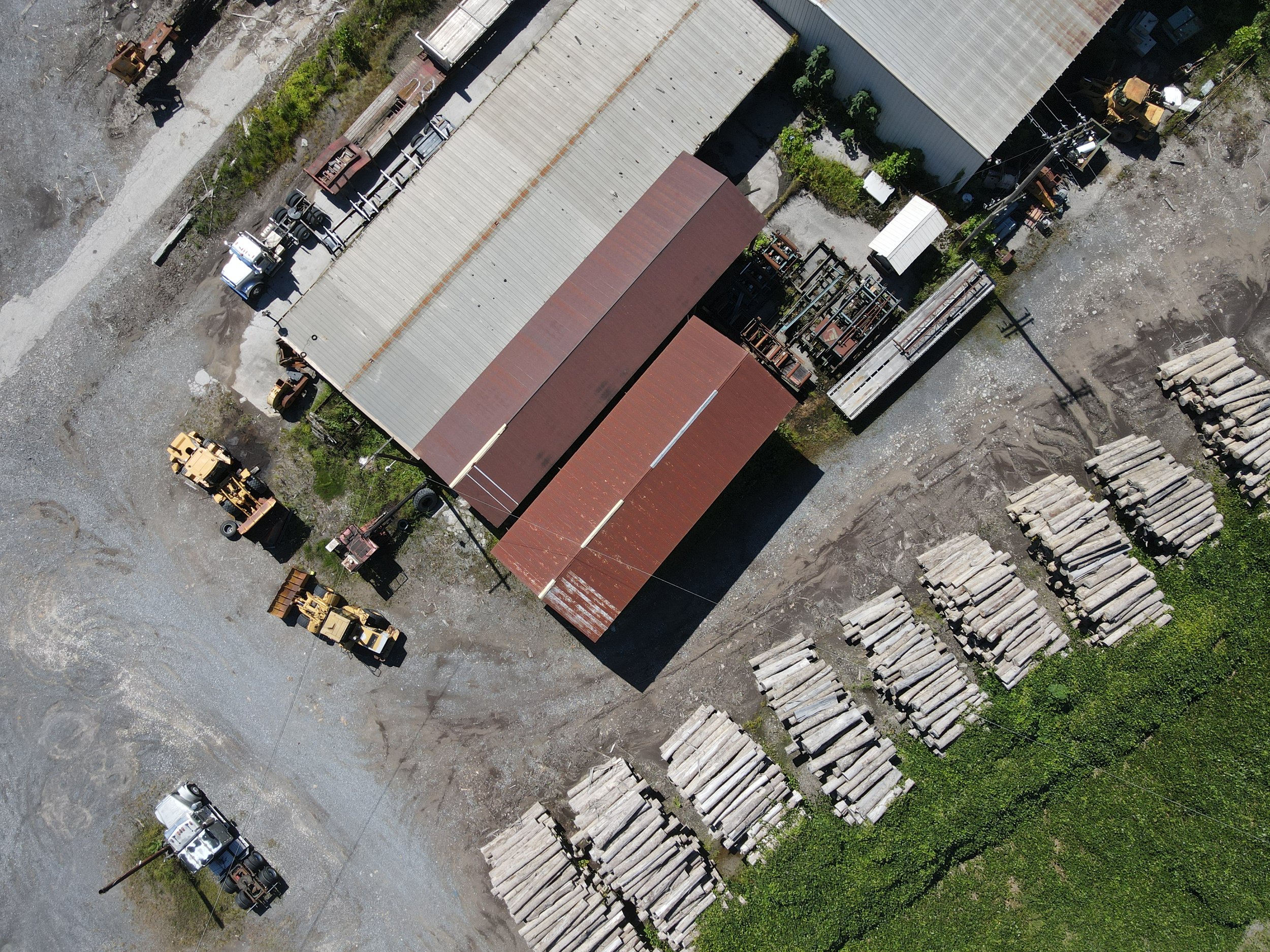 An aerial view of an industrial outdoor storage area with several trucks and stacks of logs. There is a large building with a rust-colored roof and a smaller white building nearby. Power lines cross the area, and the ground is a mixture of gravel, dirt, and paved surfaces.