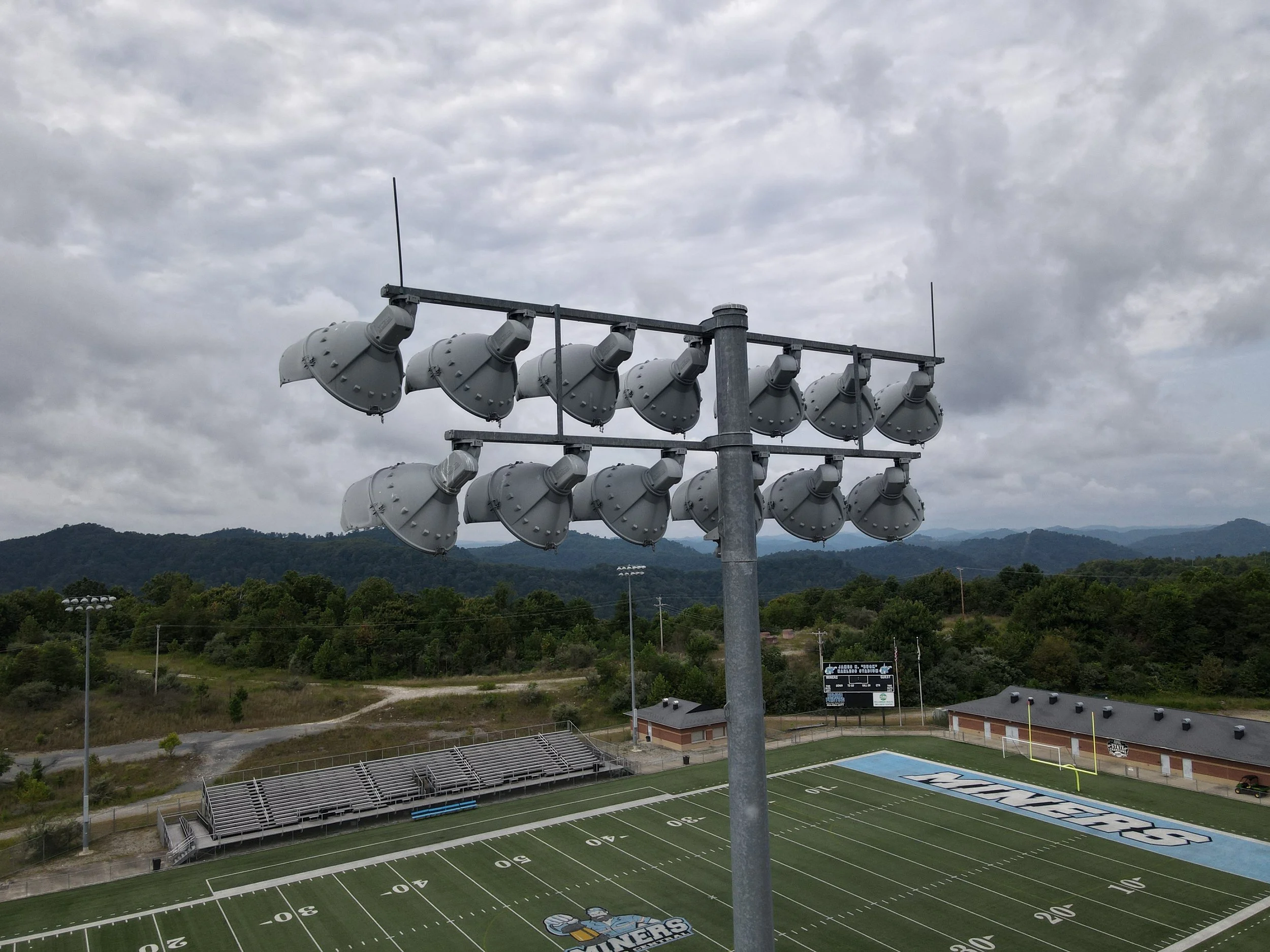A football stadium with green field and bleachers, seen from above, with a large scoreboard and cloudy sky.