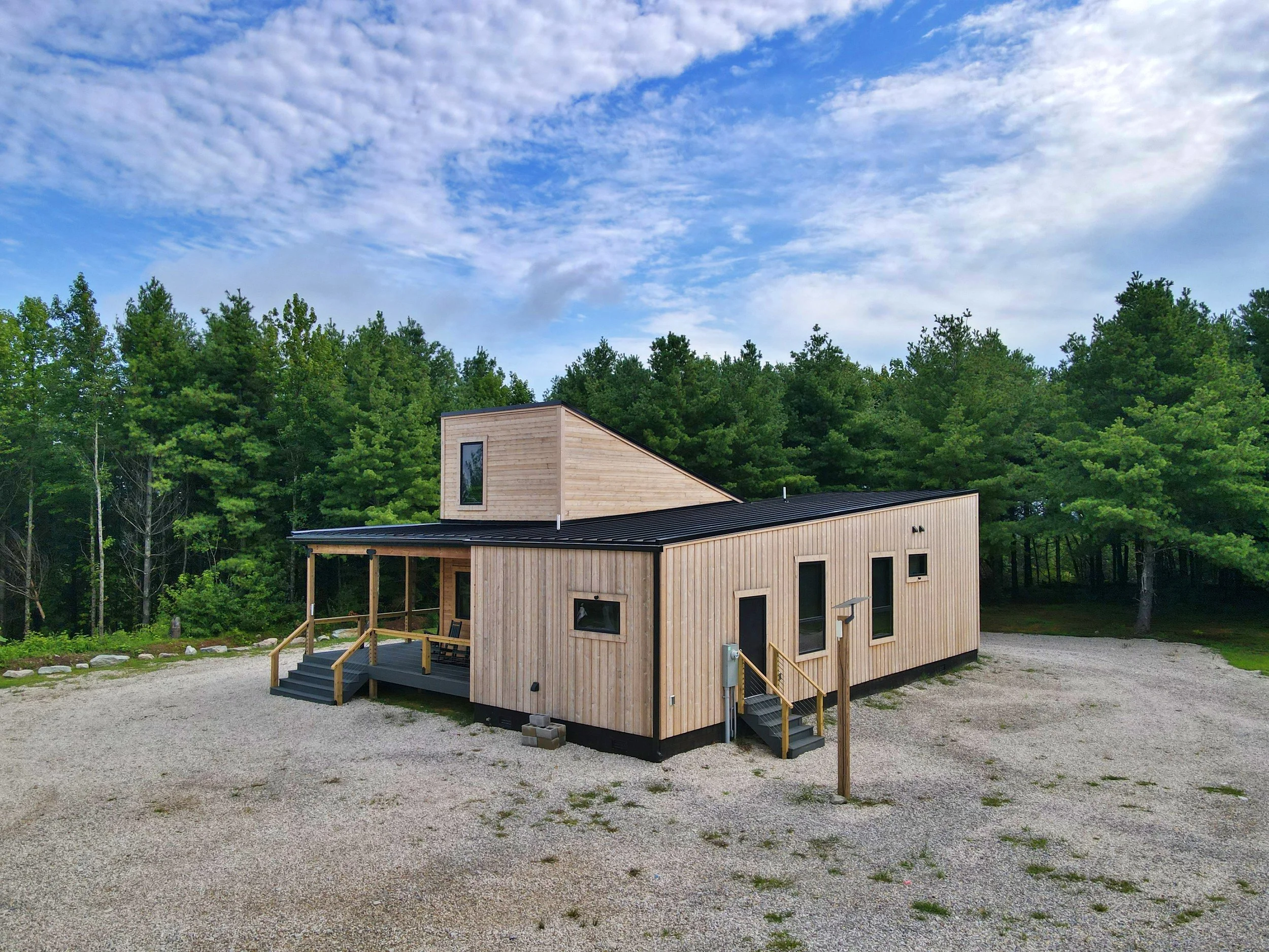 Modern wooden house with a flat roof, set in a rural area with green trees and a gravel driveway, under a partly cloudy blue sky.