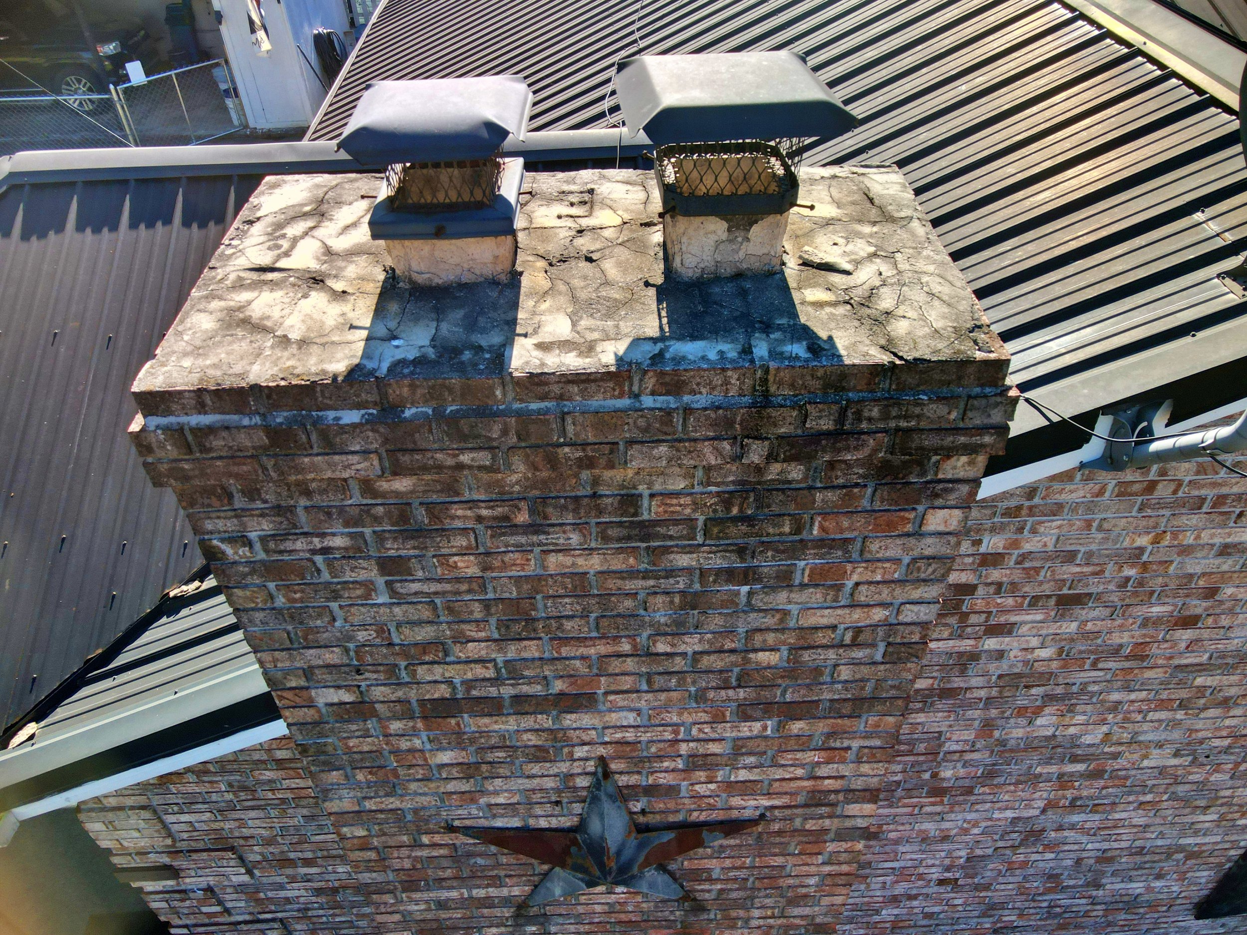 A rooftop chimney with two metal caps and a brick construction. The chimney shows signs of weathering with some cracks and discoloration. There is a metallic vent pipe extending from the chimney and a radar dish attached at the base of the chimney.