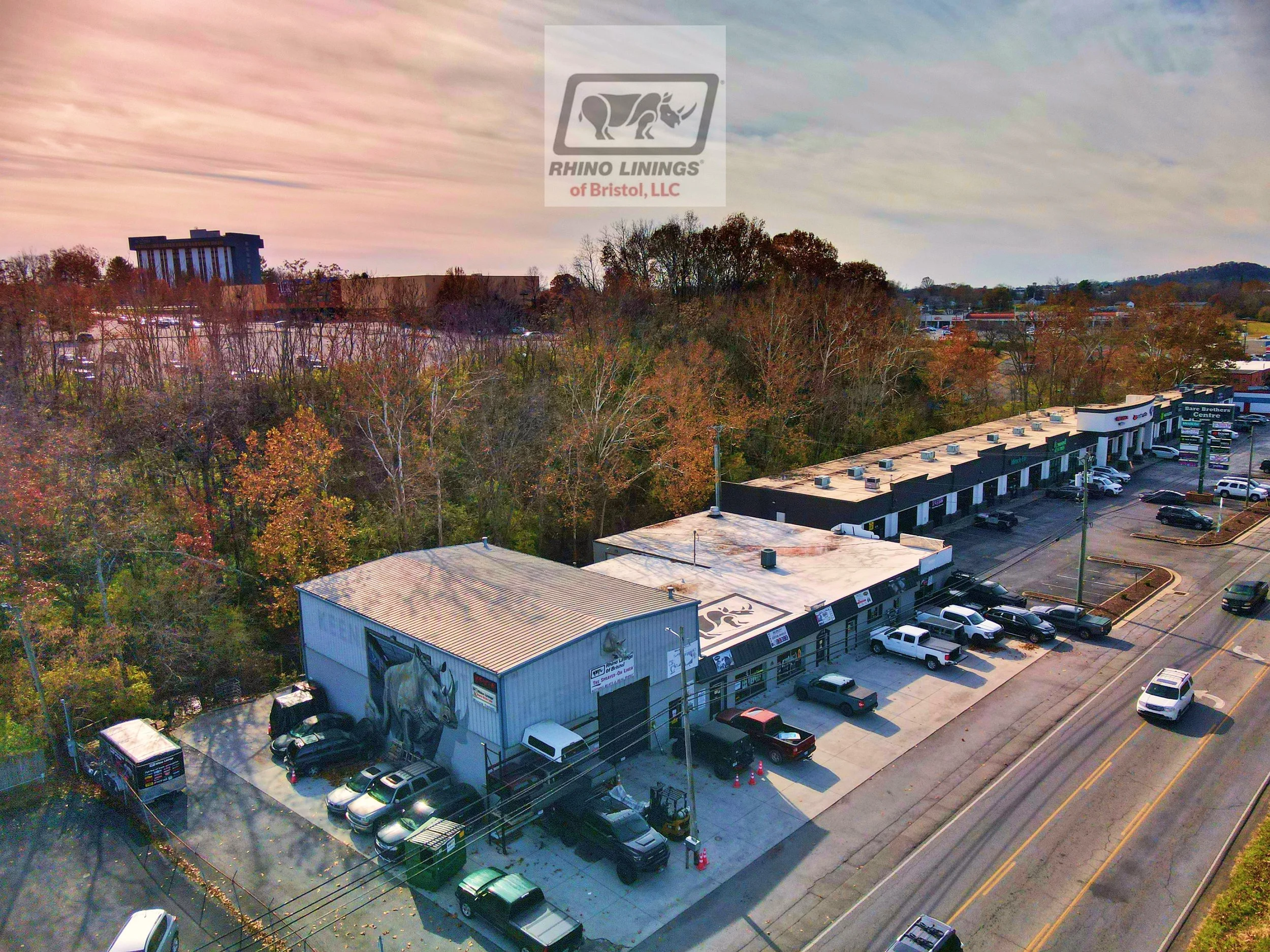 A commercial building with a parking lot filled with cars, next to a road. Behind the building, there are trees with fall foliage, and in the background, there is a small city or town with various buildings and a larger multi-story building visible.