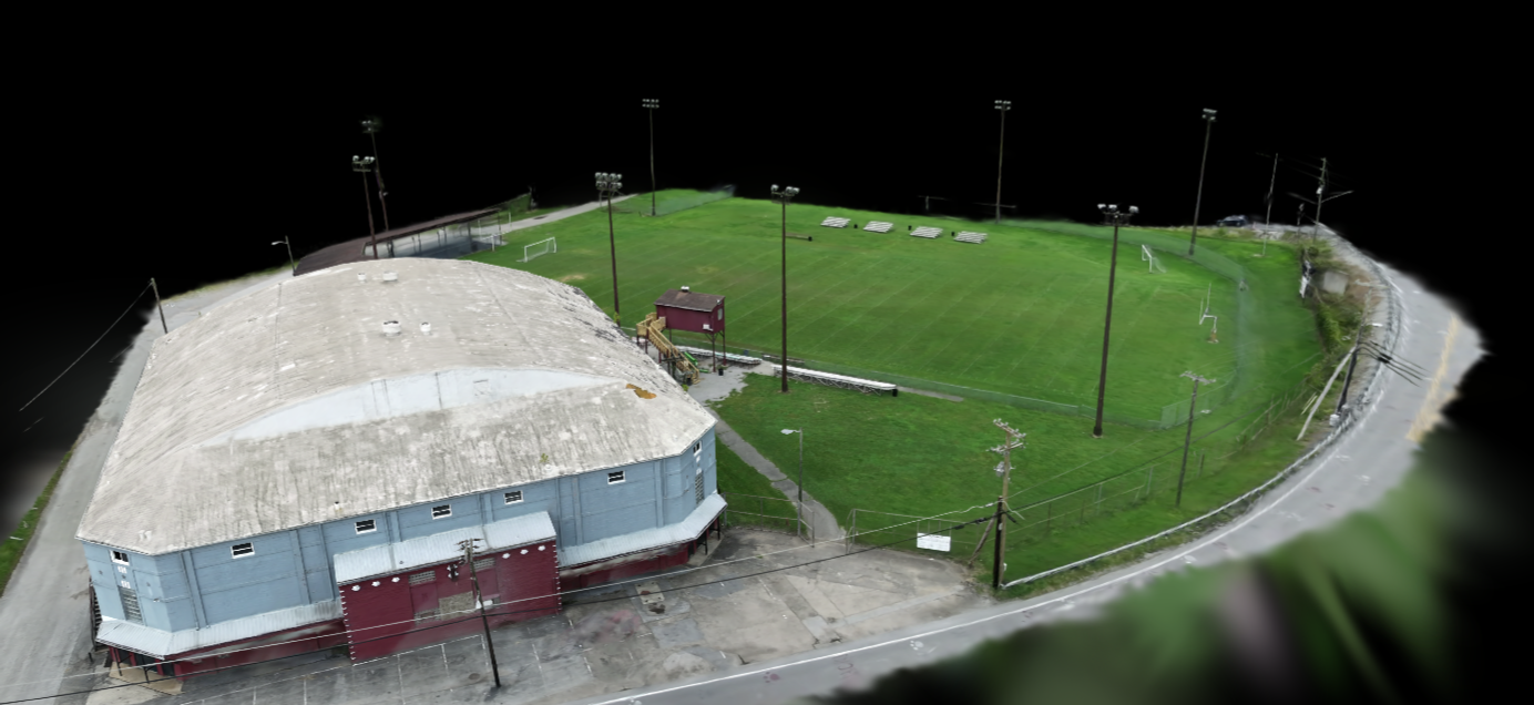 Aerial view of a small sports complex with a green soccer field, surrounded by a tall chain-link fence, and a large, older, light-blue building with a curved roof adjacent to the field.