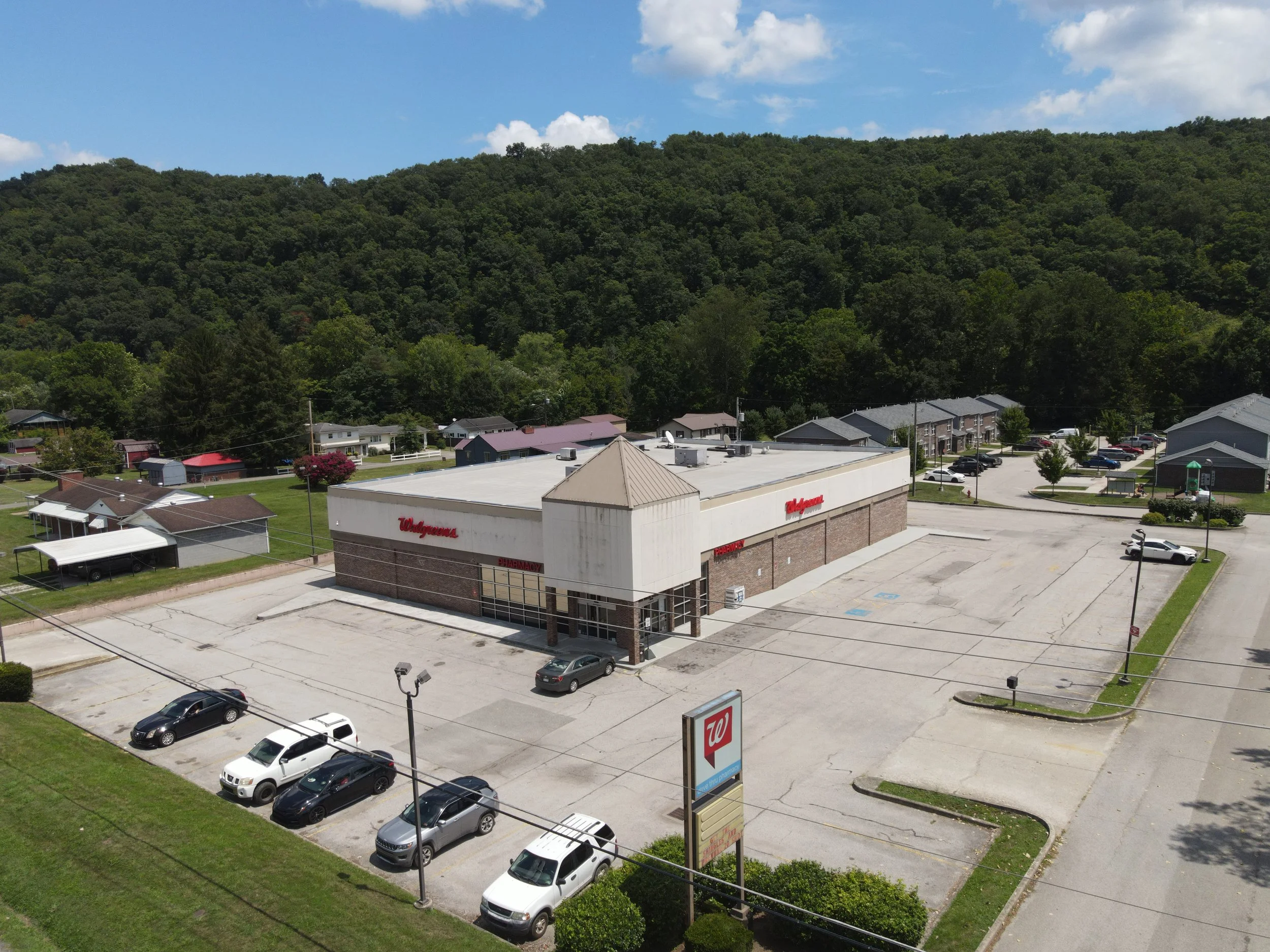 A parking lot with several cars parked near a Walgreens store, with a hillside covered in trees and blue sky with some clouds in the background.