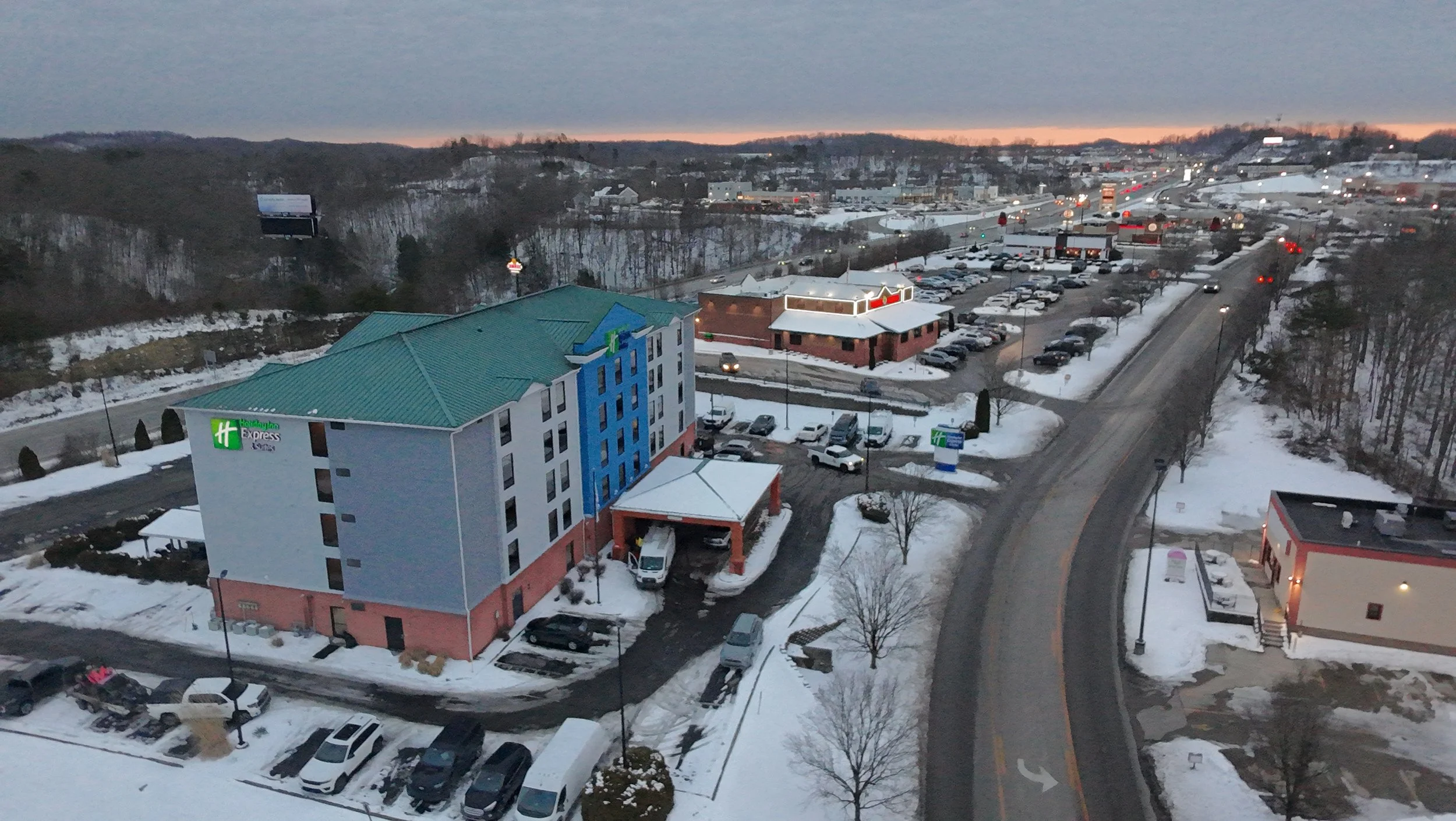 A snowy cityscape at dusk with a Holiday Inn Express hotel in the foreground, a parking lot filled with cars, and a busy road with moving vehicles. Hills and some billboards are visible in the background.