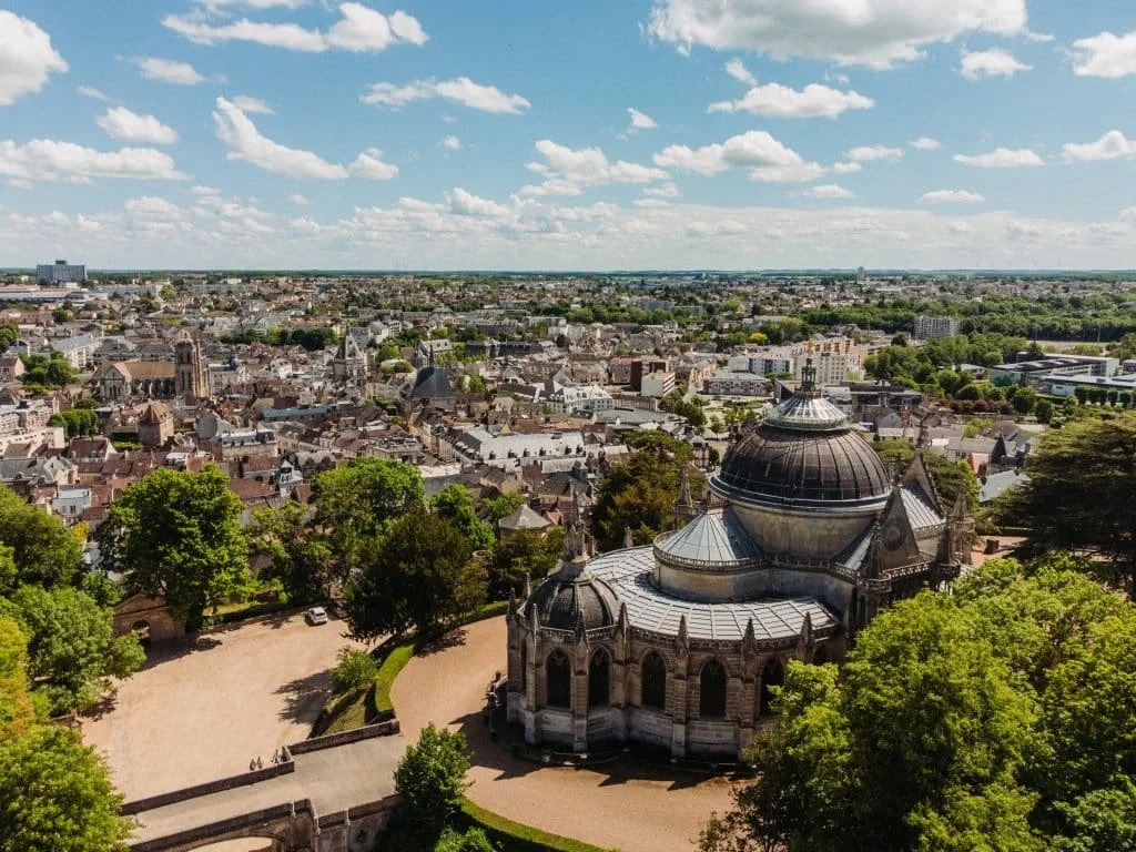 Une vue aérienne d'une ville avec une grande église gothique au premier plan, entourée d'arbres et de bâtiments résidentiels, sous un ciel bleu avec quelques nuages.