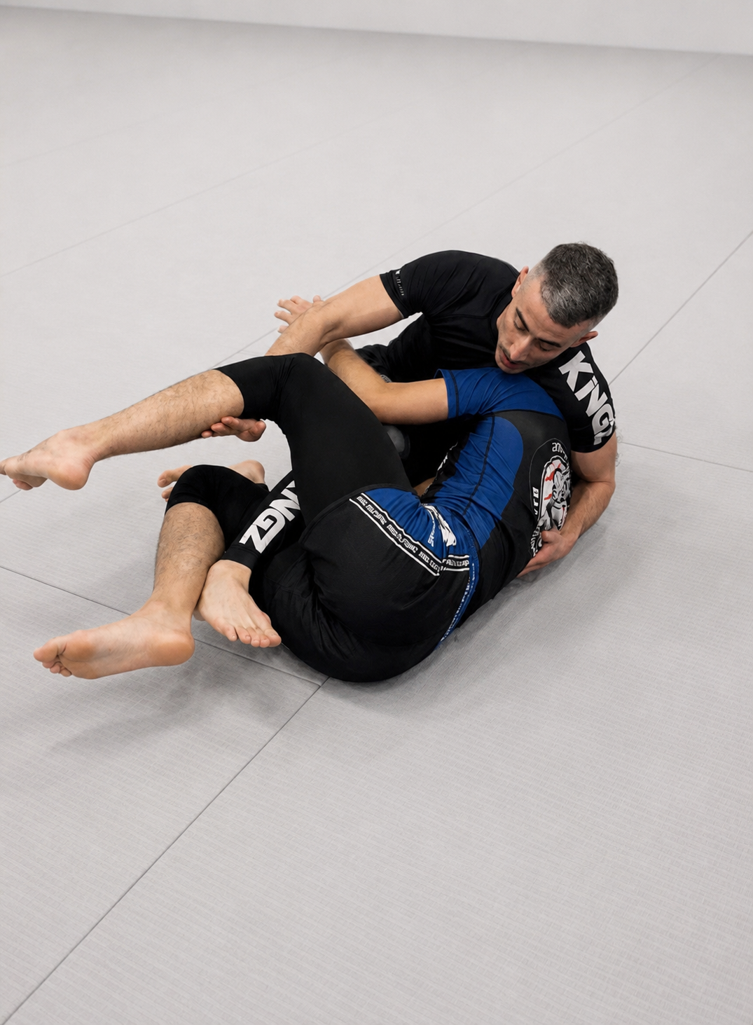 Deux hommes pratiquant le jiu-jitsu brésilien sur un tapis d'entraînement, dont l'un en position de contrôle sur l'autre.