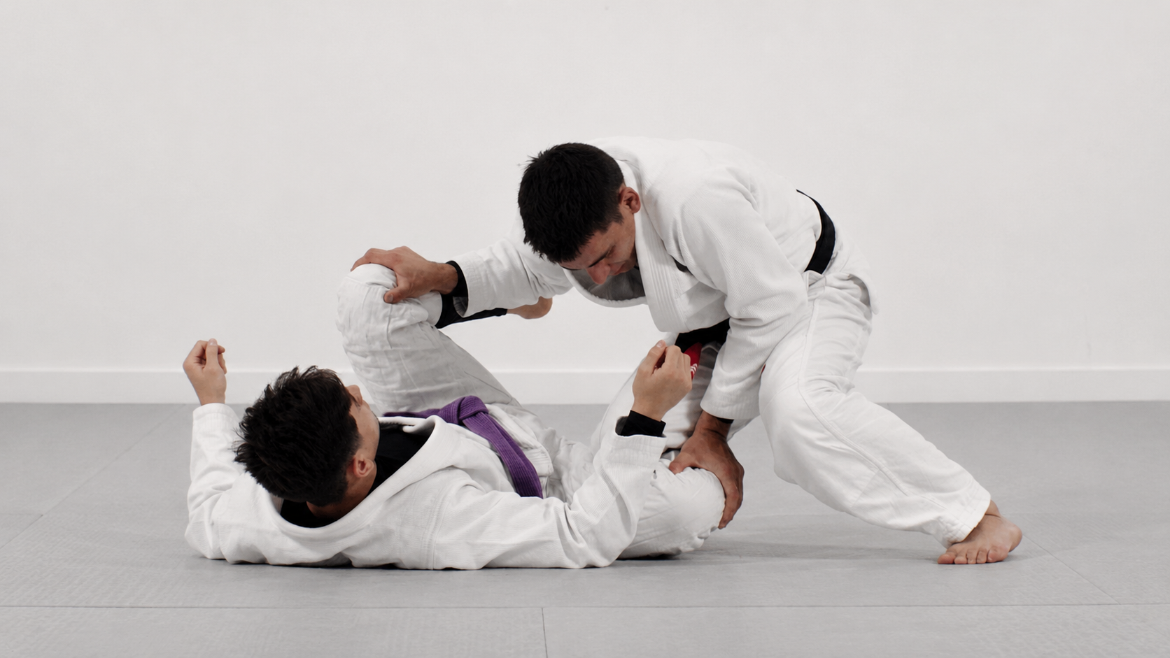 Deux pratiquants de judo en uniforme blanc, l'un au sol avec une ceinture violette, l'autre en position de contrôle, sur un tapis de judo, dans une salle d'entraînement.