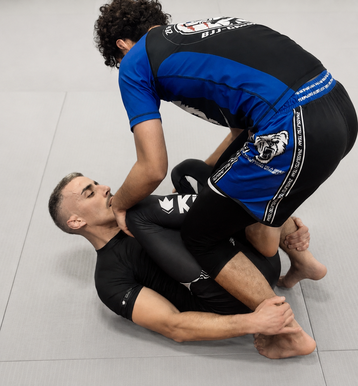 Deux hommes s'entraînent au jiu-jitsu brésilien sur un tapis gris, l'un en position de contrôle, l'autre essayant de défendre.