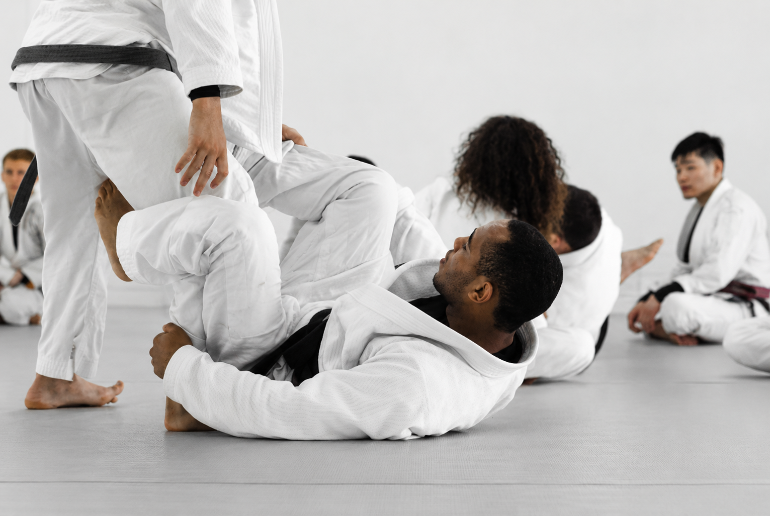 Des pratiquants de jjb en kimono blanc exécutent une technique sur le tapis d'un dojo, d'autres observateurs en kimono blanc sont assis au fond.
