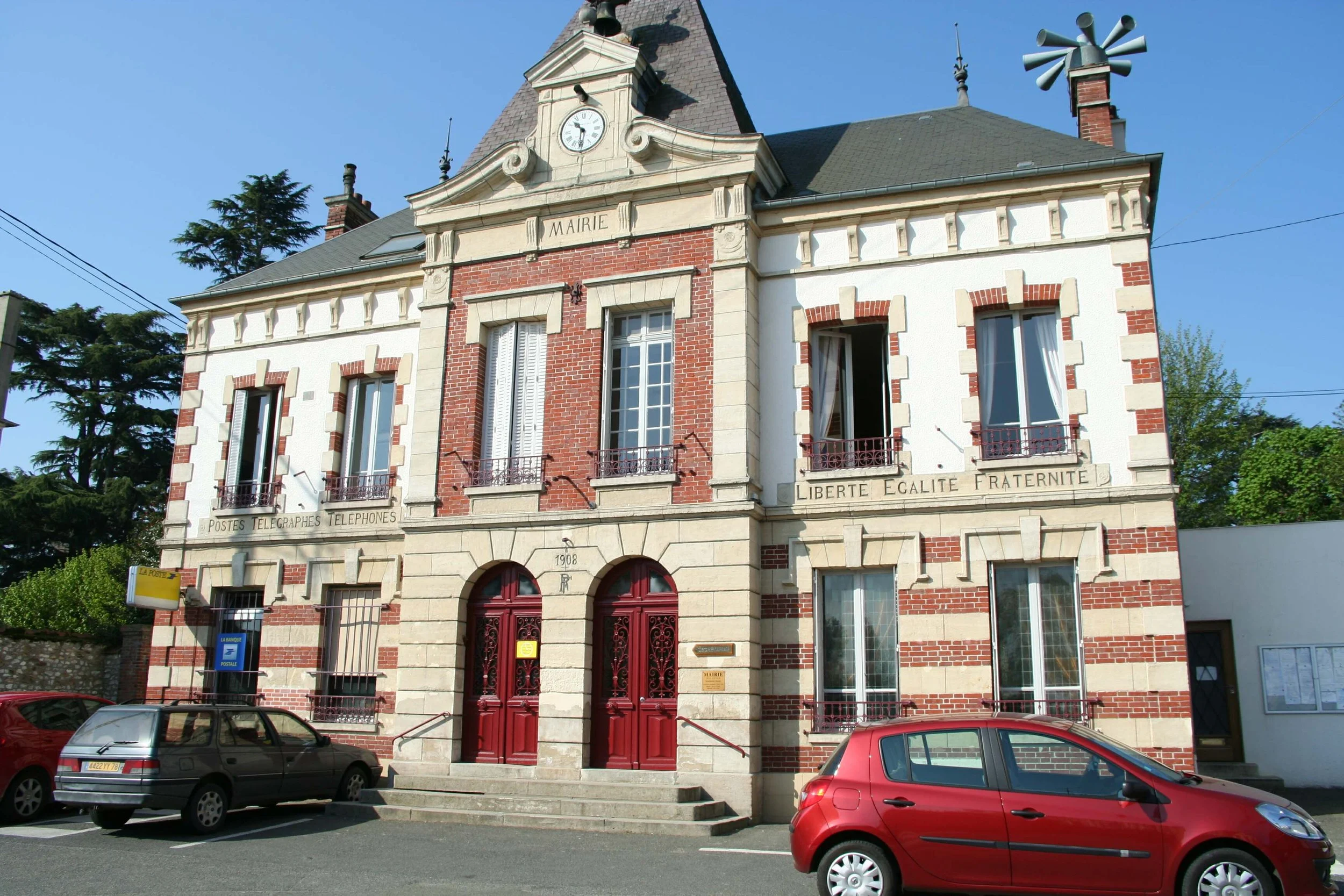 Bâtiment de style classique avec une façade en briques rouges et détails en pierre, portant une inscription 'MAIRIE'. Il a de grandes fenêtres, une porte en bois rouge ornée de décorations en métal, et se trouve en face d'un parking avec des voitures rouges et grises. Le bâtiment a un horloge en haut avec une toiture en pente et plusieurs éléments décoratifs en métal. Il y a une inscription 'LIBERTÉ ÉGALITÉ FRATERNITÉ' sous une corniche.