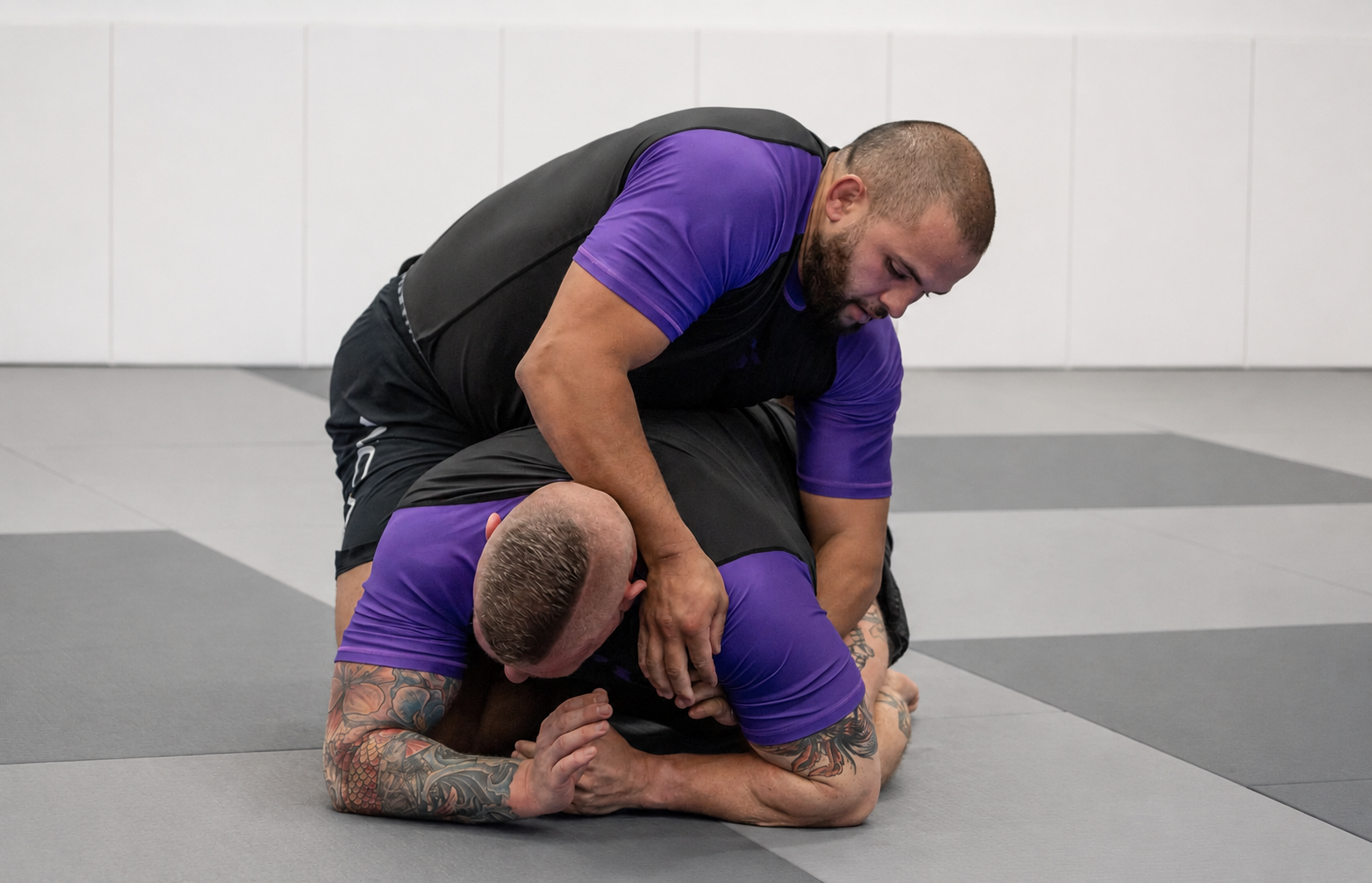 Deux hommes pratiquant le jiu-jitsu dans une salle d'entraînement, l'un au sol, l'autre en position de contrôle, tous deux portant des vêtements de sport violets et noirs.