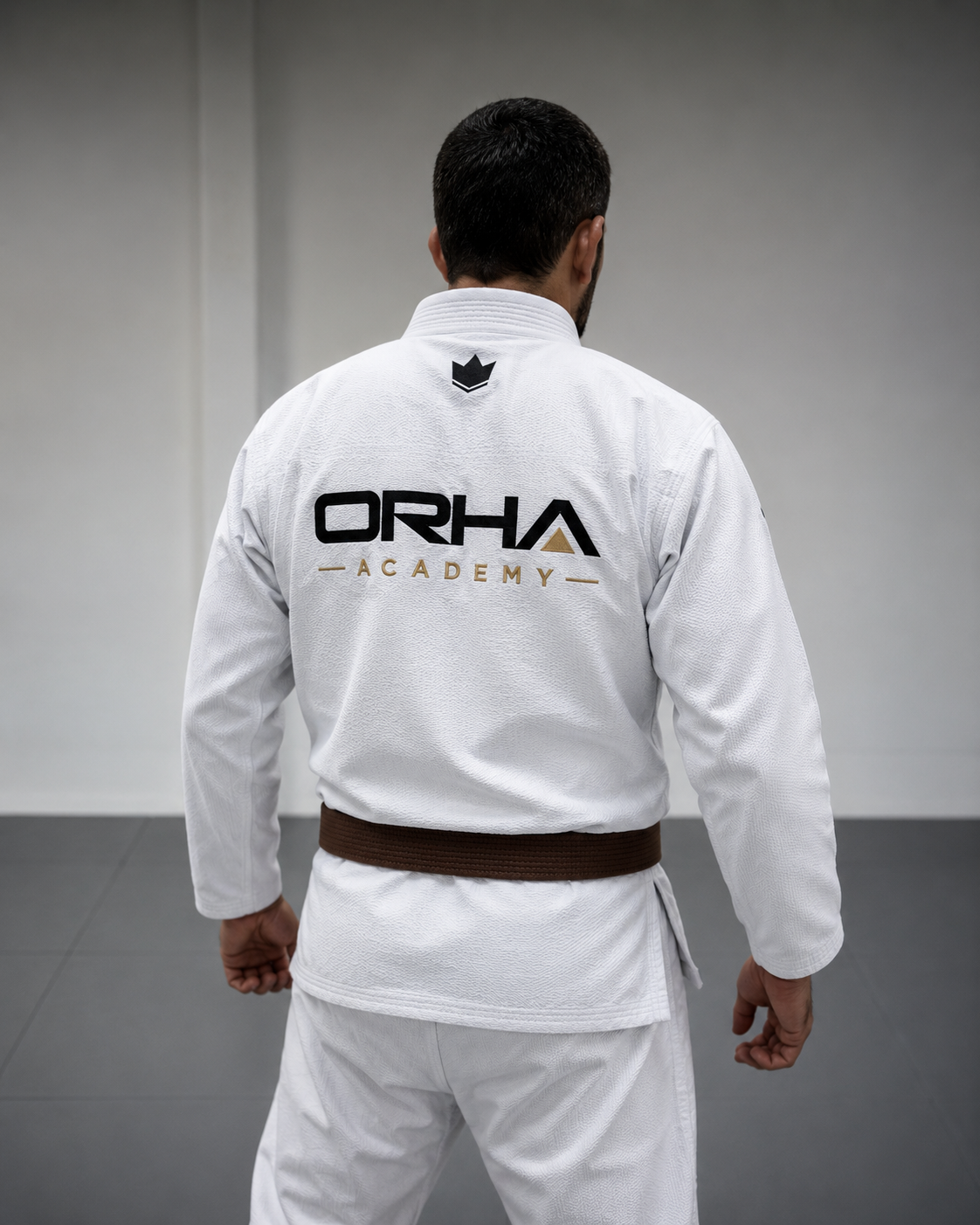 Un pratiquant de jjb vêtu d'une kimono blanche avec un ceinture brune, vu de dos, dans un dojo, portant un logo Orha Academy sur le dos.