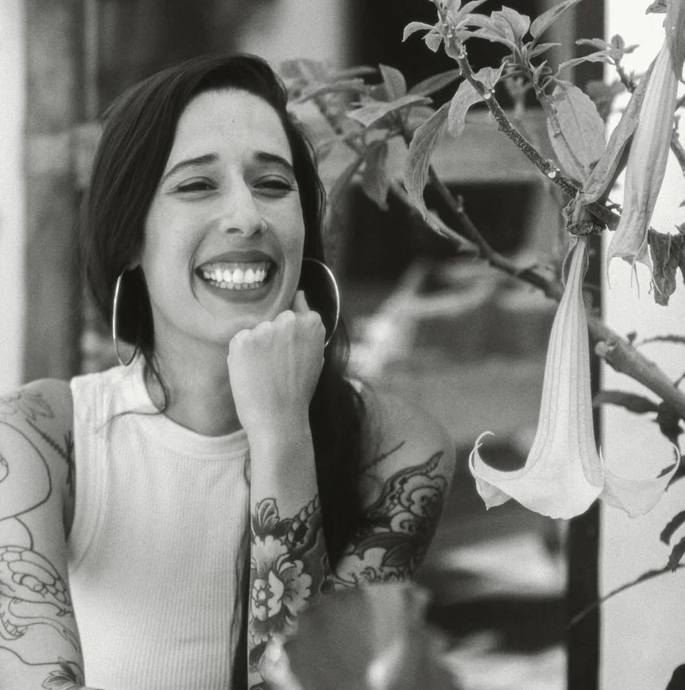 A woman smiling and resting her chin on her hand, with tattoos on her arms, wearing hoop earrings, in front of a large hanging flower in a black and white photo.