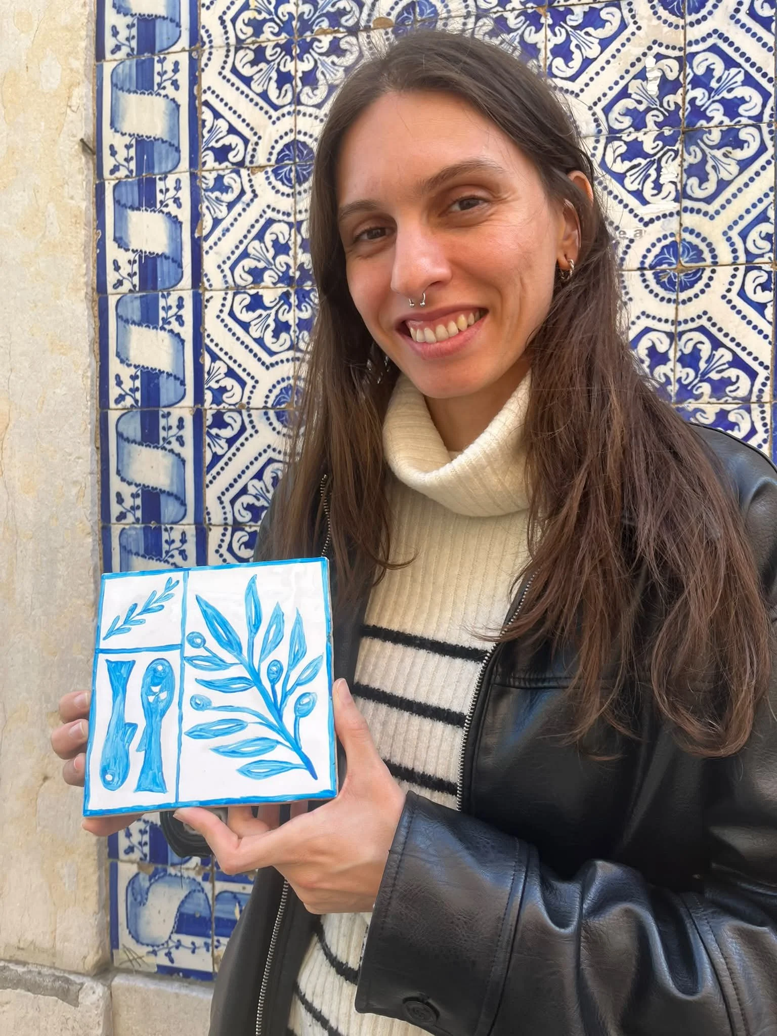 A woman smiling and holding a blue and white tile art piece with leaf and olive branch designs, standing in front of a decorative blue and white tiled wall.