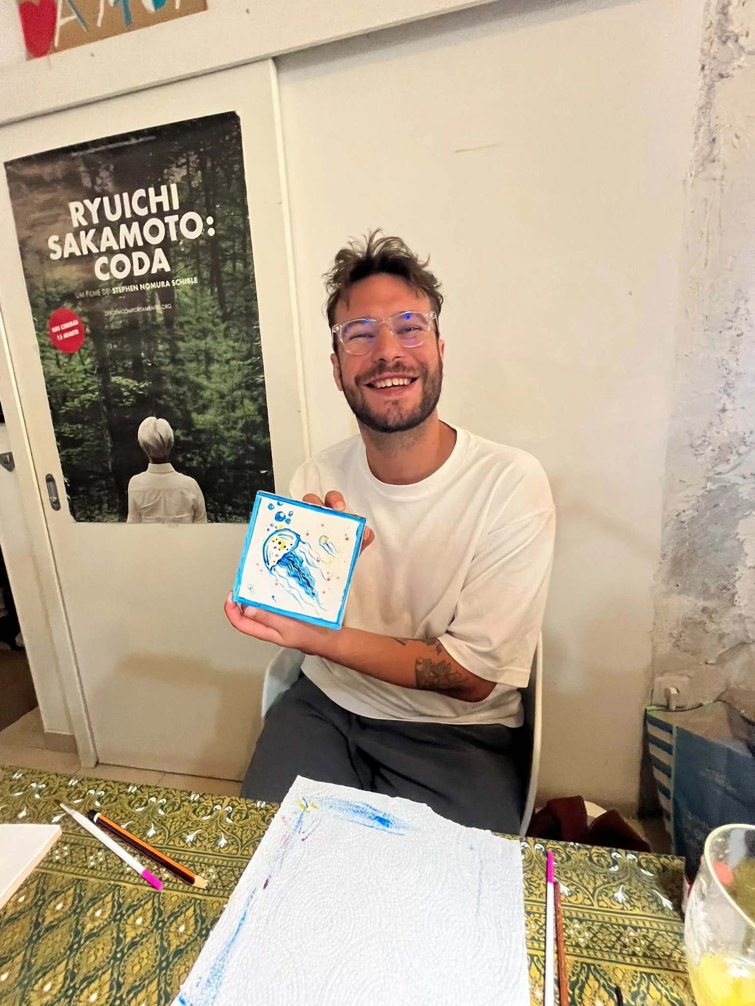 A man with glasses and a beard smiling while holding a colorful drawing of a rainbow and water. He is sitting at a table with art supplies, with a banner and a poster in the background.