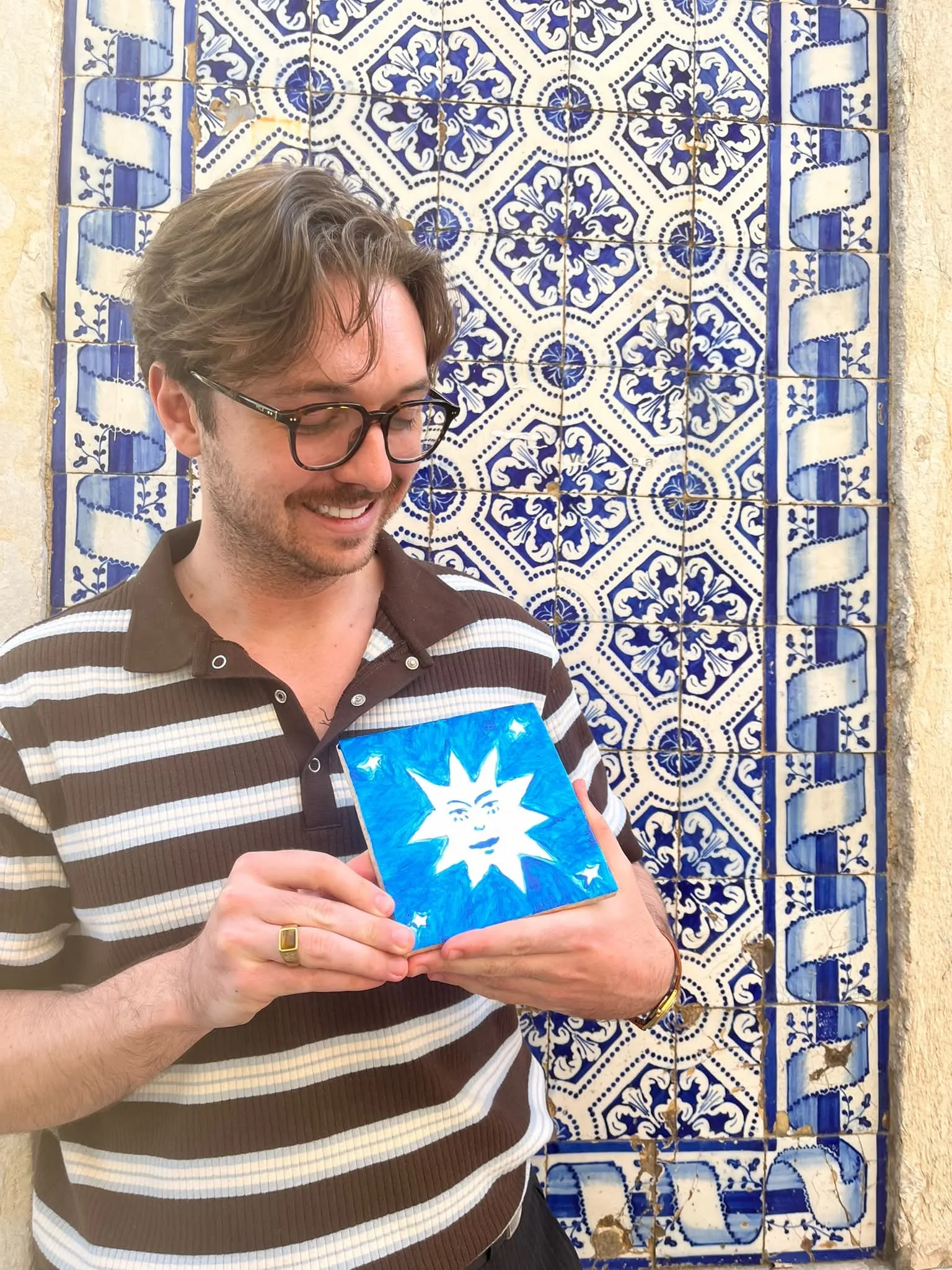 A young man wearing glasses and a striped polo shirt is standing in front of a patterned blue and white tile wall. He is smiling and holding a small blue piece of art with a face and stars on it.