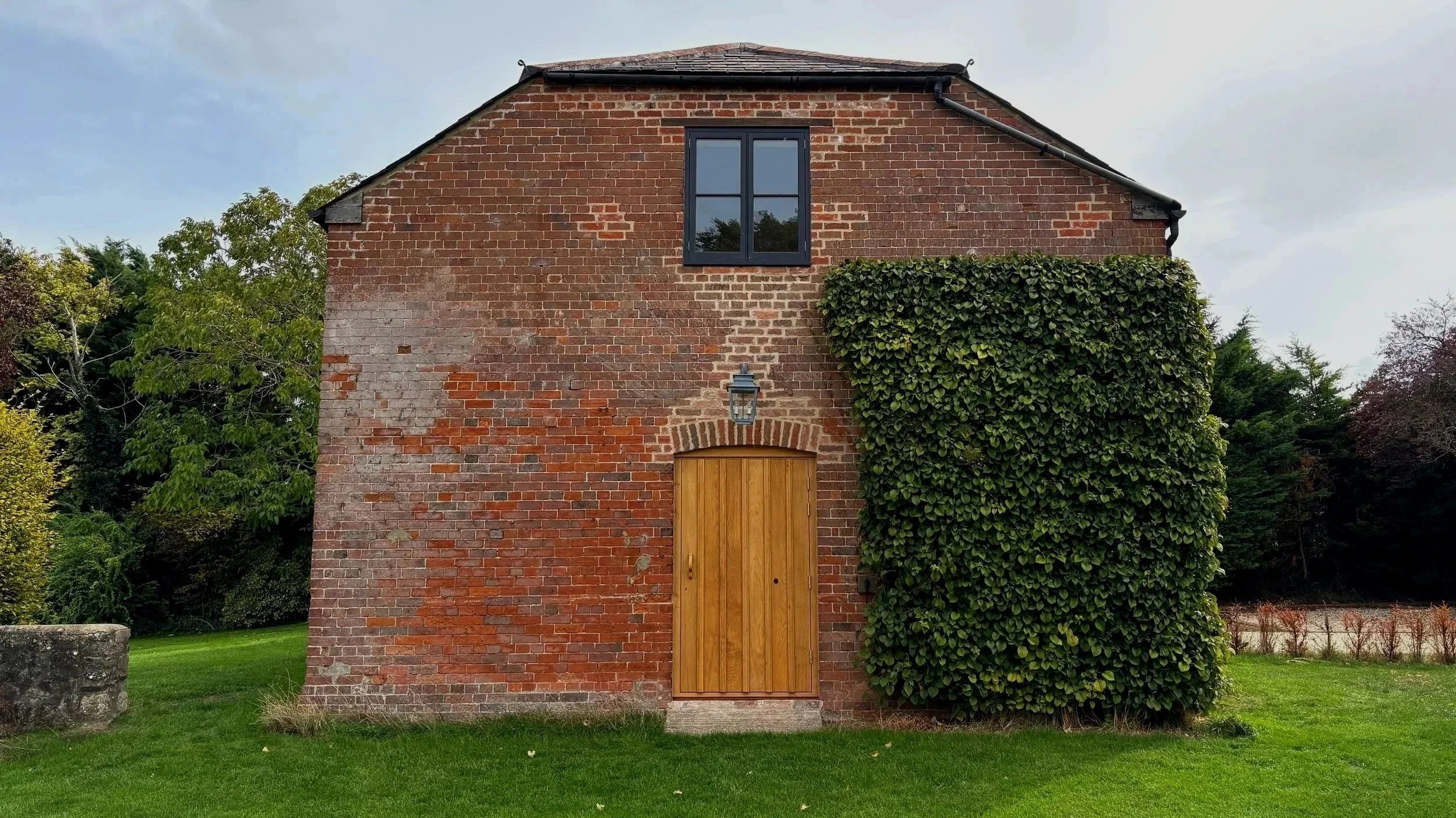Red brick building with a wooden door, a black-framed window, and green ivy growing on the right side, surrounded by green lawn and trees.