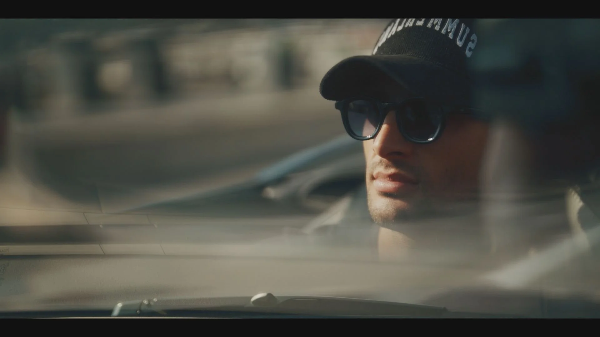 A man with glasses and a baseball cap seen through a car windshield.