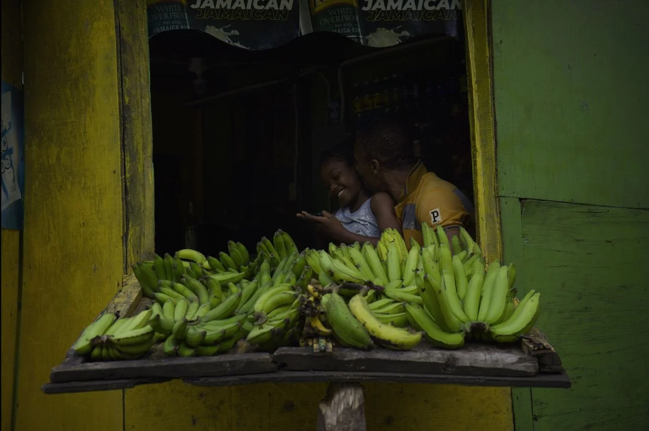 A girl and a man smiling and looking at a phone at a banana stand with bananas displayed in front of them. The stand has yellow and green wooden walls.