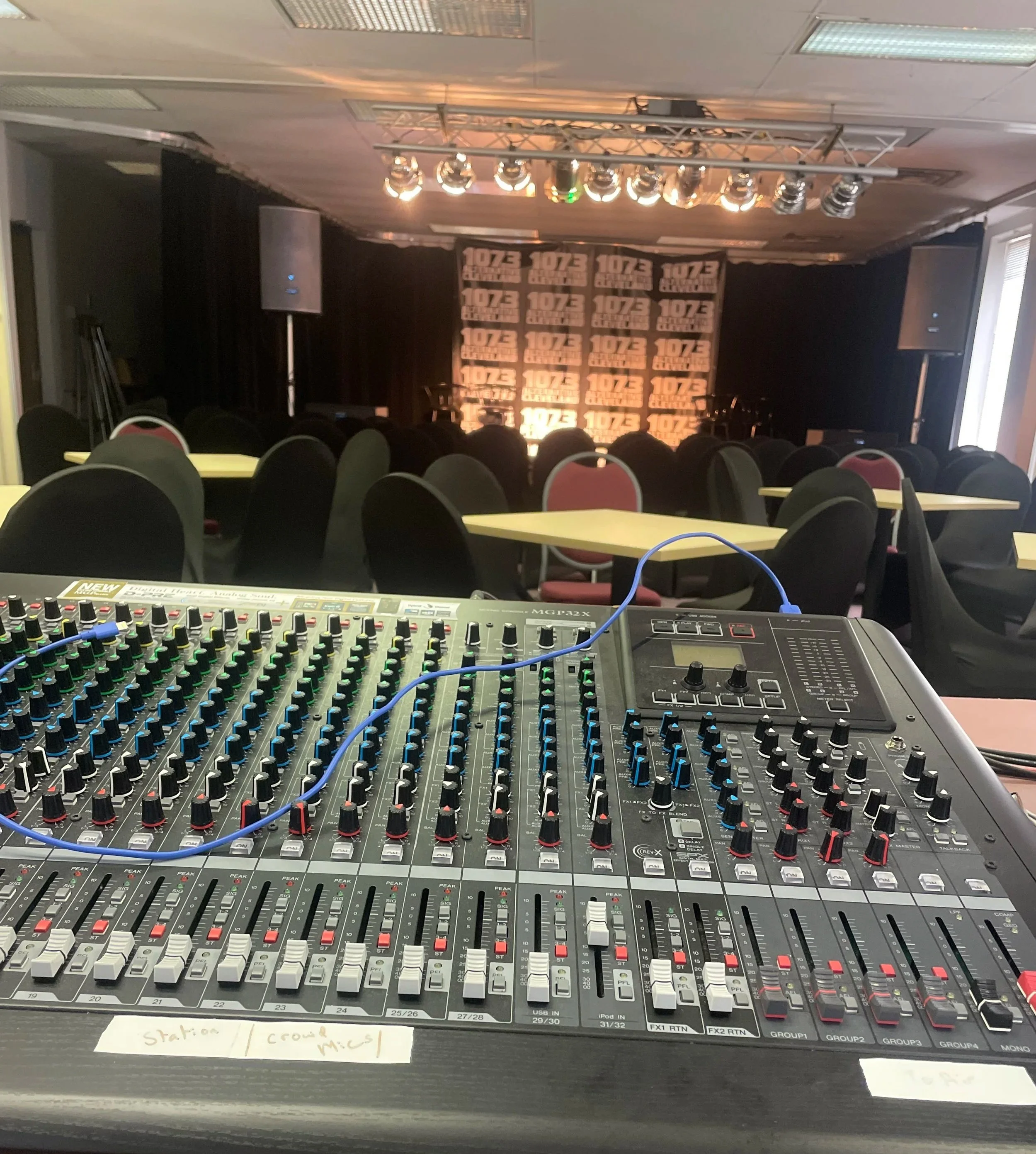 Sound mixing console, stage setup with speakers and a backdrop featuring the logo '107.3 ALPHA' in a dimly lit room, with tables and chairs in the foreground.