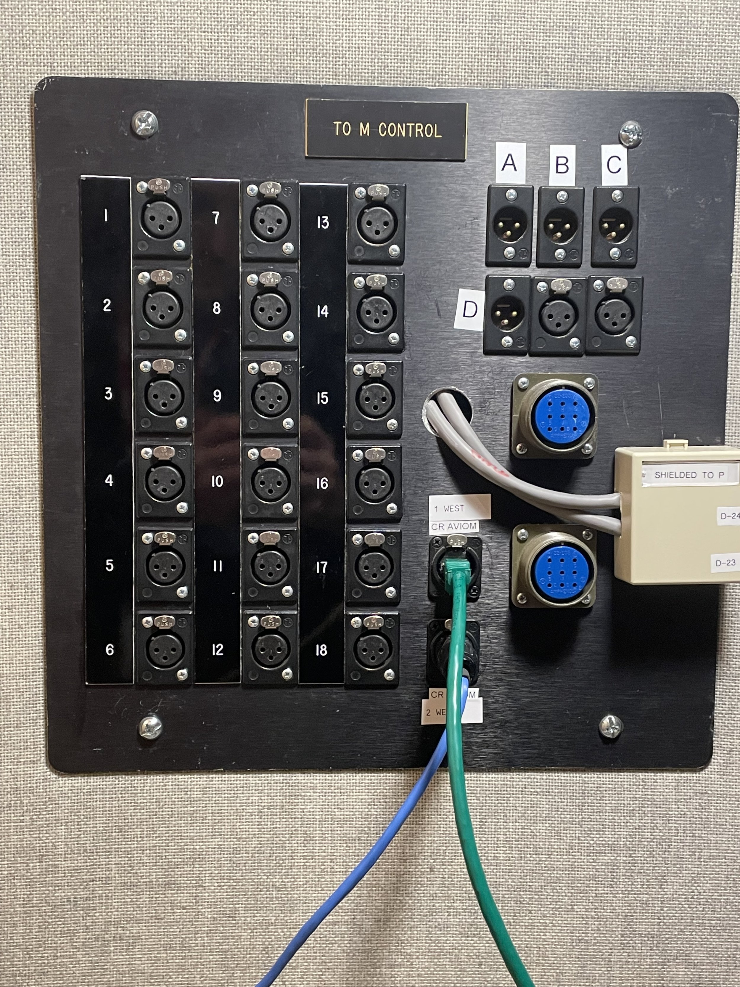 A wall-mounted control panel with multiple labeled sections including numbered sockets, ports, and a cable connected. It has labels such as 'TO M CONTROL', 'A', 'B', 'C', 'D', and various numbers from 1 to 18. There are also some handwritten labels and cables plugged into different ports.