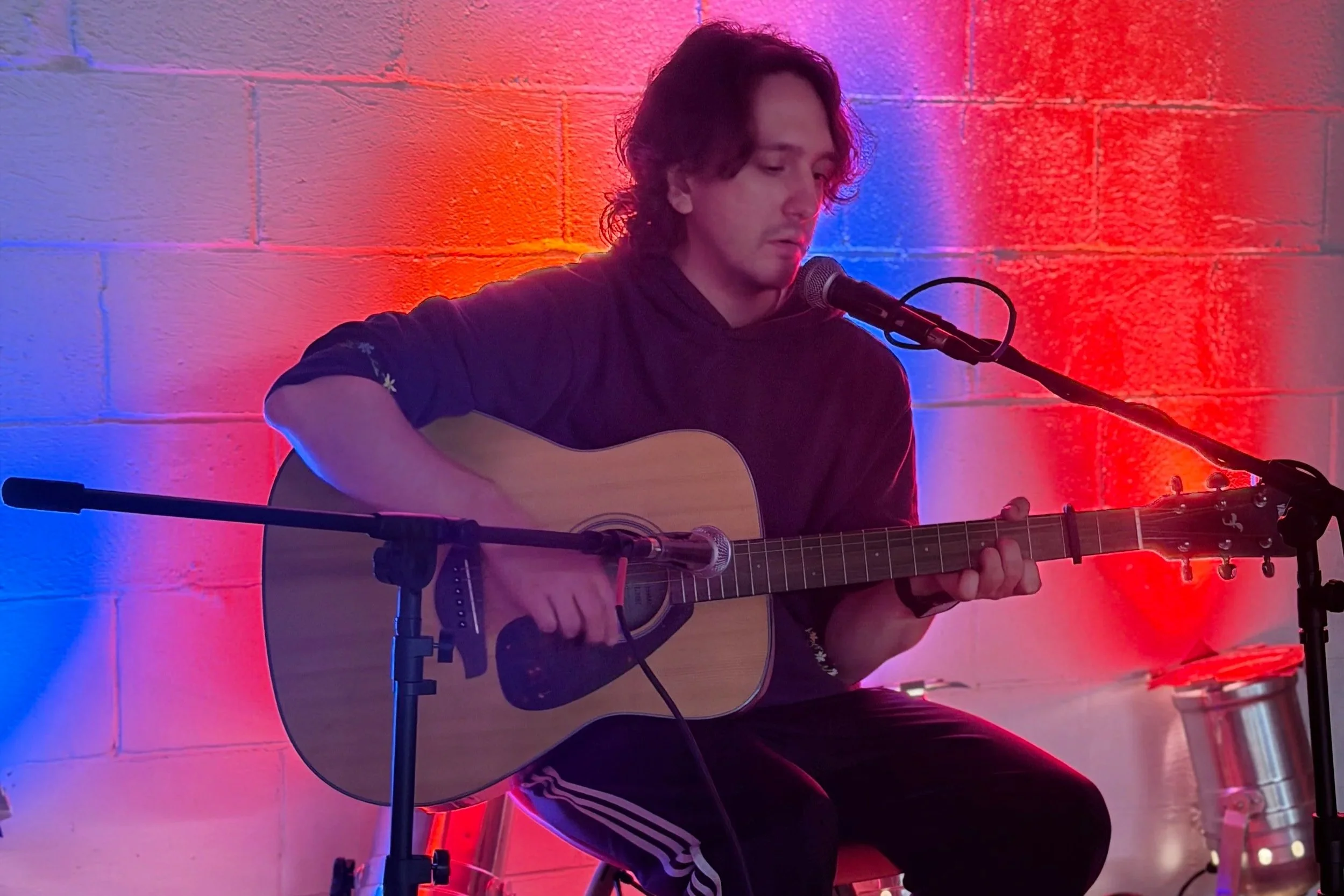 A person with long dark hair, wearing a dark hoodie with floral details on the sleeve, playing an acoustic guitar and singing into a microphone. The background has a colorful lighting effect with red, blue, and purple hues on a brick wall.