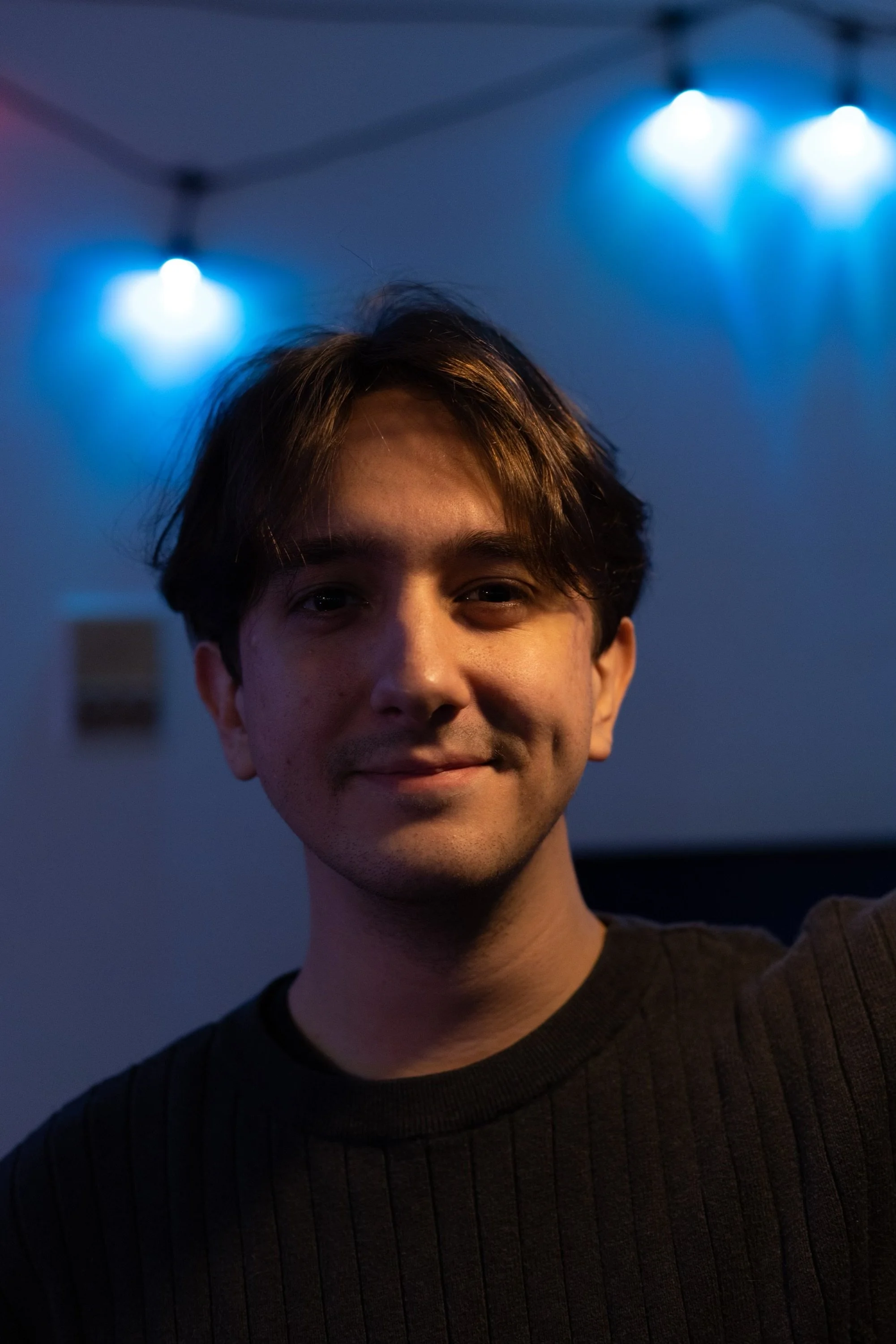 A young man with brown hair and a faint smile, wearing a black ribbed sweater, standing indoors with blue and white lighting in the background.