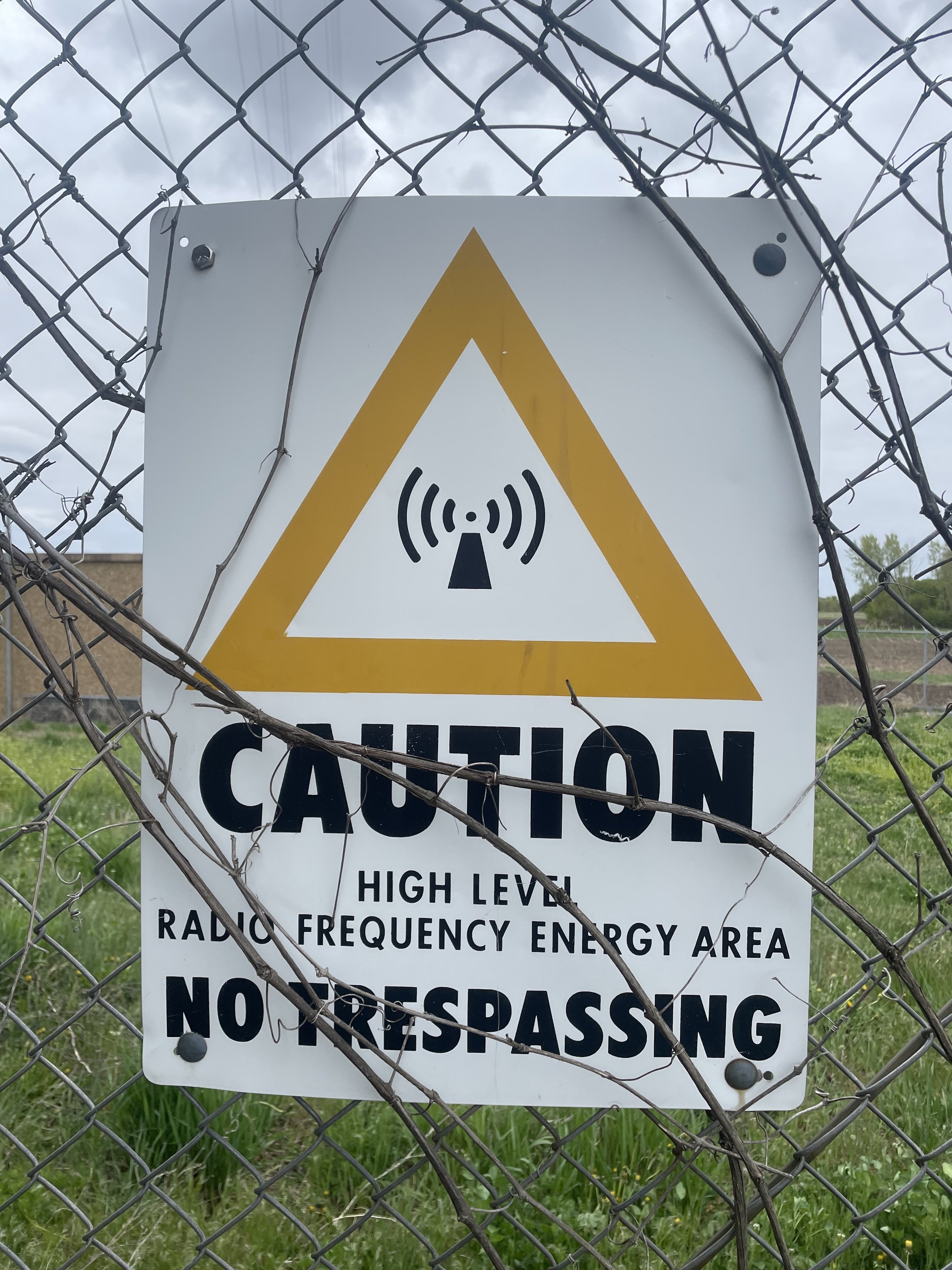 A warning sign on a chain-link fence indicating a high voltage range area, with a skull and crossbones and radiating wave symbols, and additional text warning no trespassing.