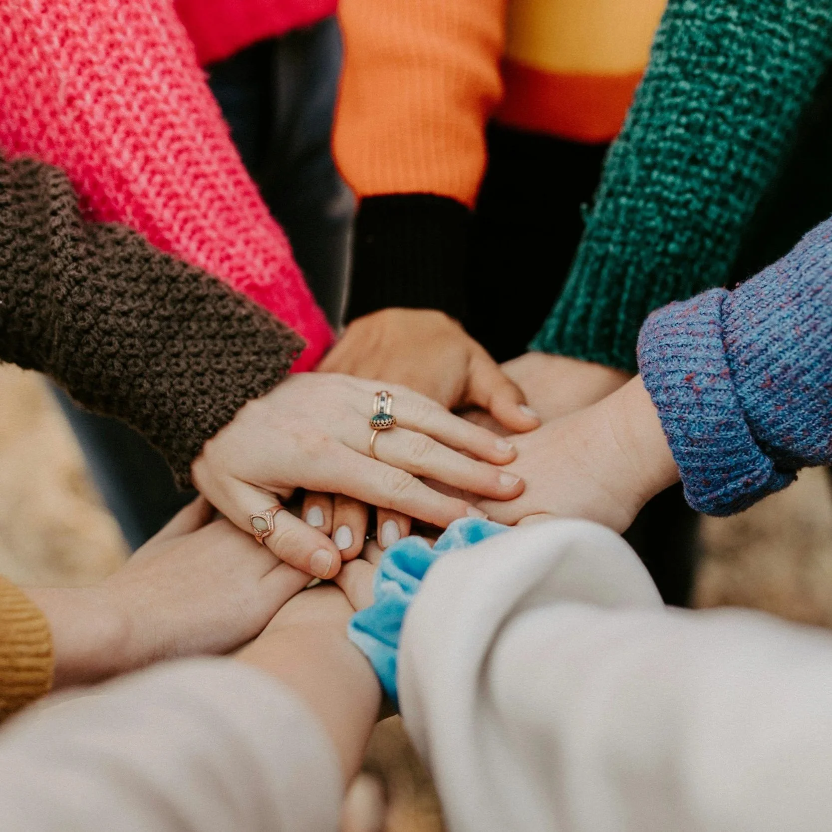 Multiple hands stack together, showing a gesture of unity and support, some wearing rings. The hands are of different skin tones and are surrounded by colorful, cozy sweaters.