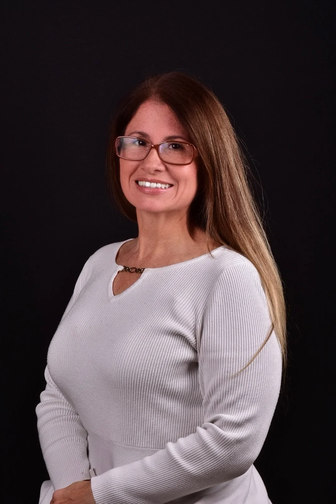 A woman with glasses, long brown hair, wearing a white ribbed sweater, smiling, posed against a black background.