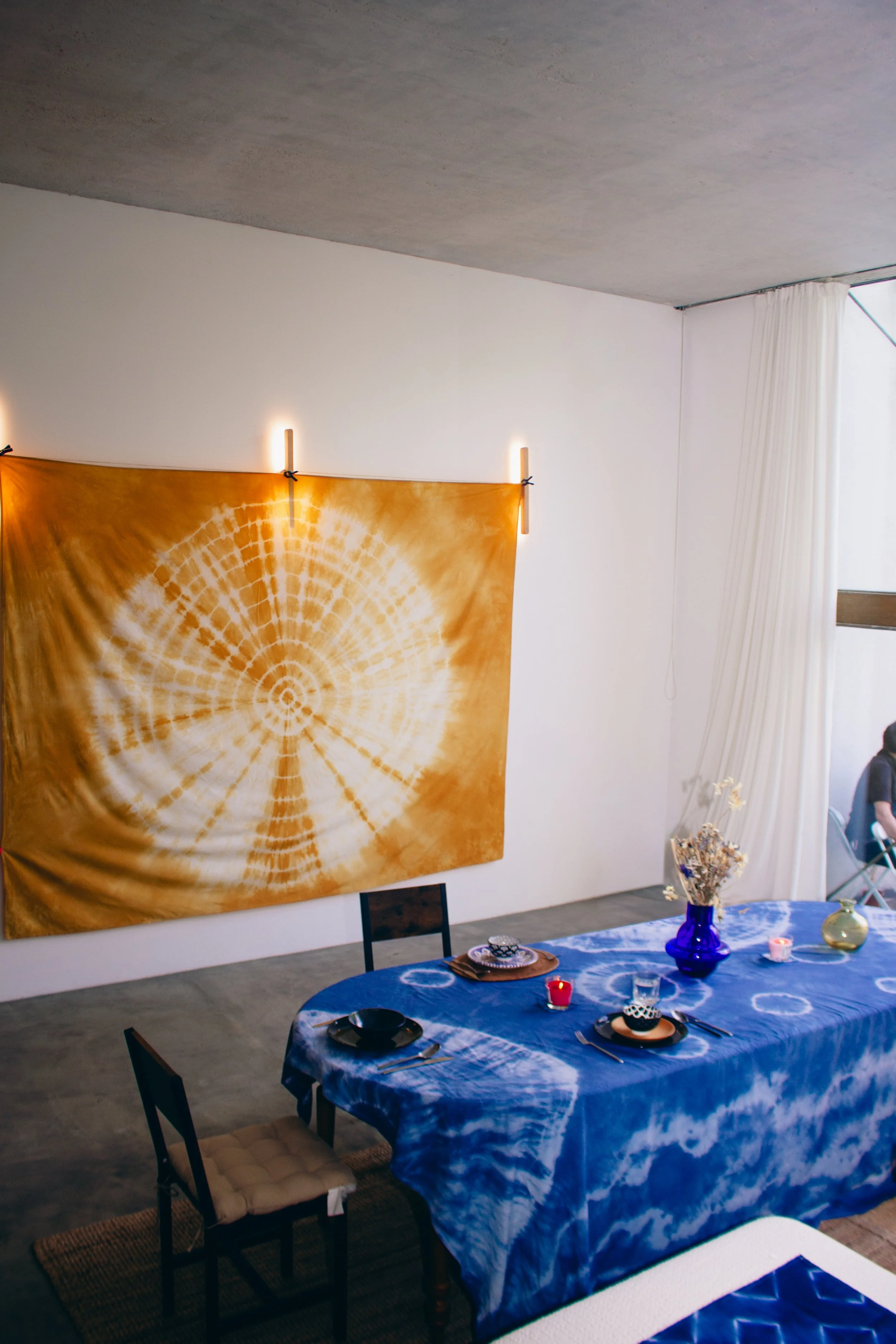 Une table dressée avec une nappe bleue en shibori et des assiettes, entourée de chaises, dans une pièce décorée avec une grande tapisserie shibori orange et des luminaires sur le mur.