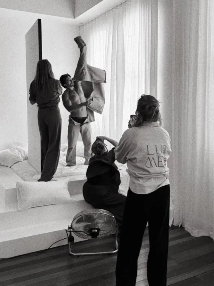 A man in boxers is jumping onto a bed while a woman watches, two others are seated on the bed, one appears to be taking a photo. The scene is in a bright room with curtains, a fan on the floor, and someone wearing a 'LUX MED' t-shirt.