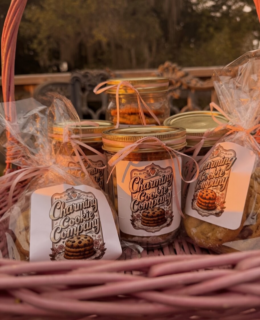 Glass jars and bags of cookies labeled 'Charming Cookie Company' displayed in a pink wicker basket outdoors.