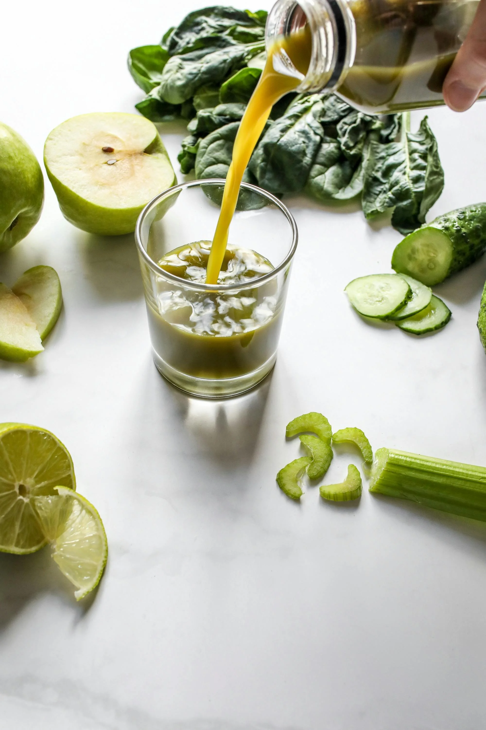 A glass of green juice being poured from a jar, surrounded by various fresh green ingredients including cucumber, apple, lemon, and leafy greens on a white surface.