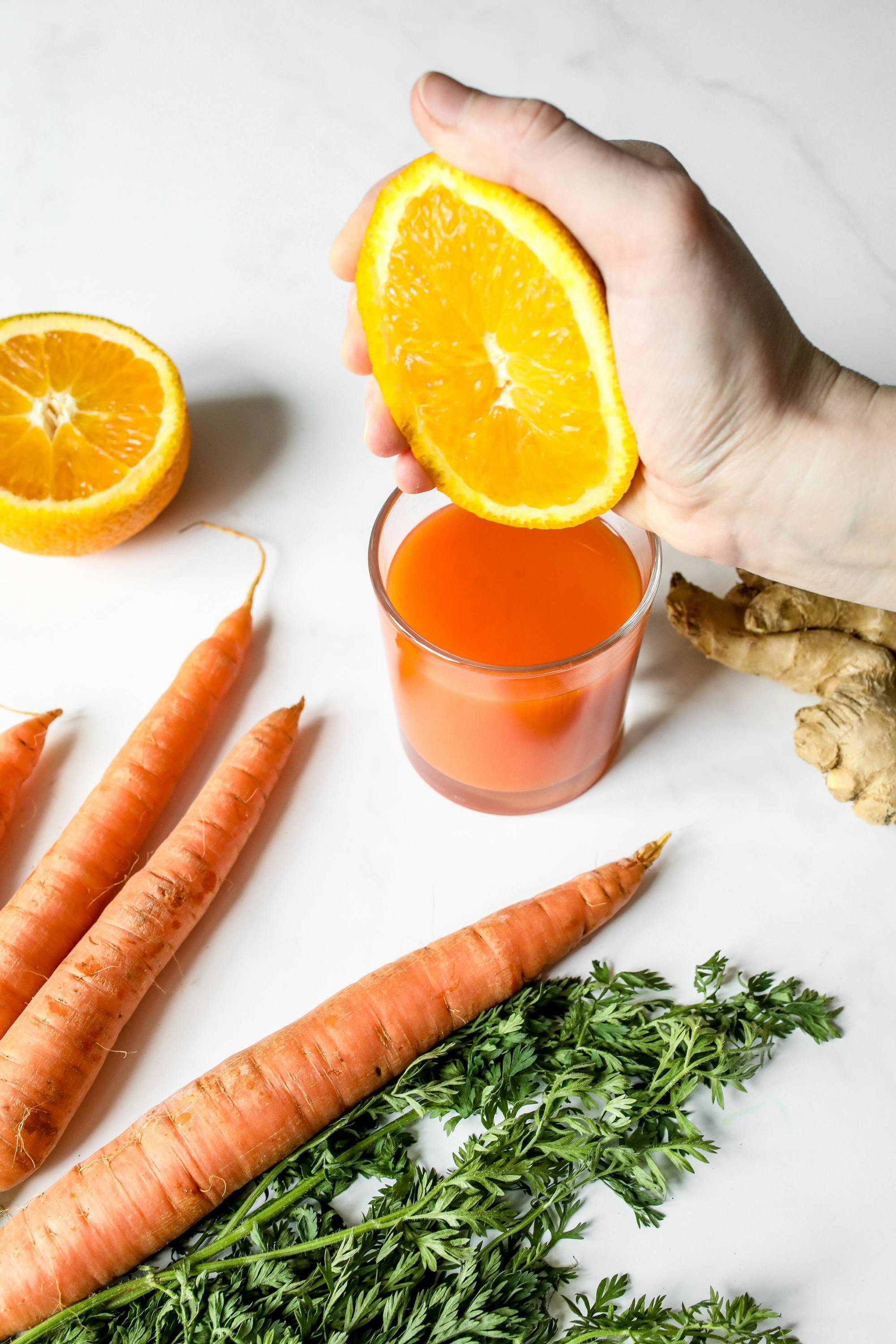 A hand holding a half orange over a glass of orange juice, with another half orange, carrots, ginger, and fresh herbs on a white surface.