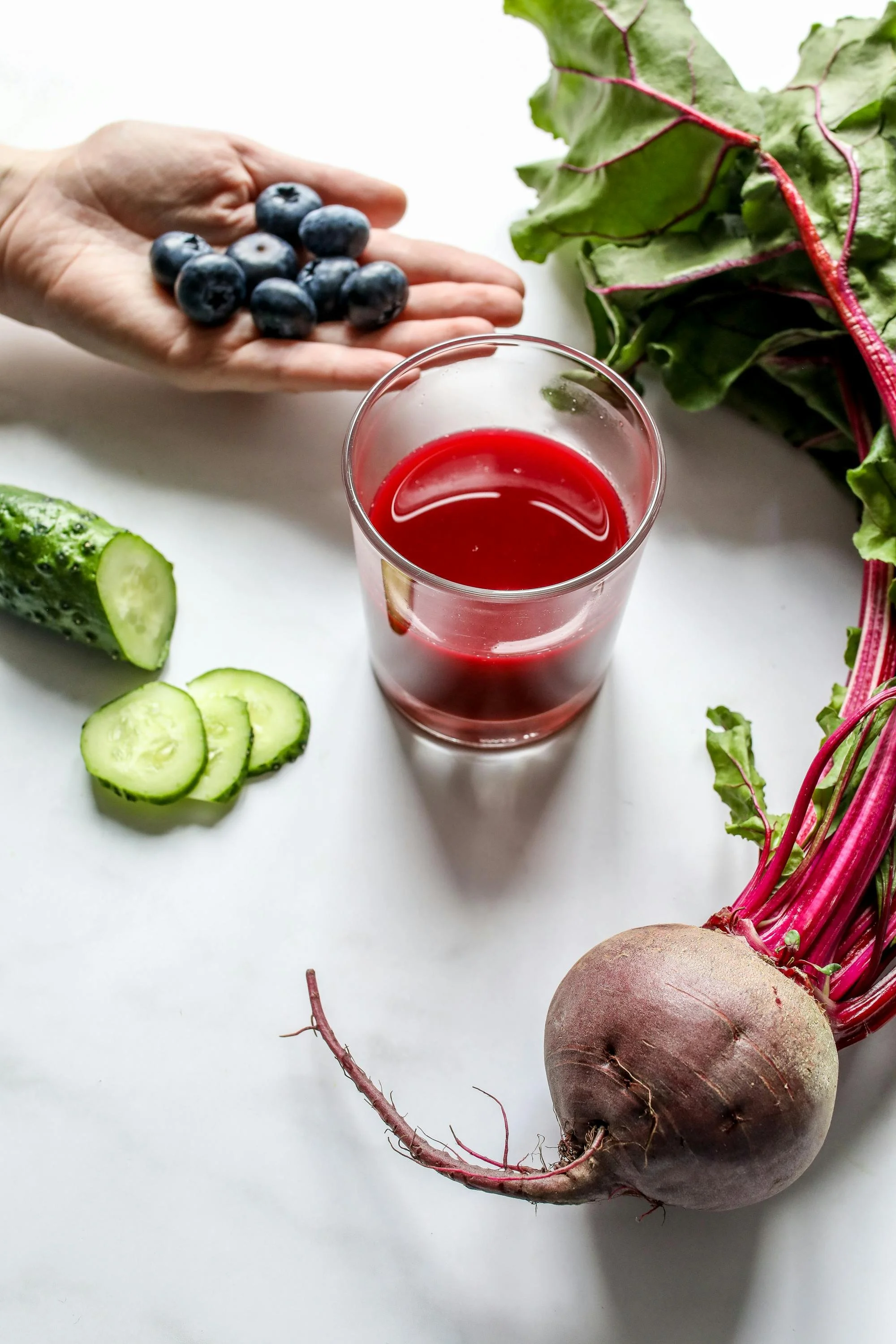A hand holding blueberries next to slices of cucumber, with Swiss chard leaves and a root vegetable on a white background.