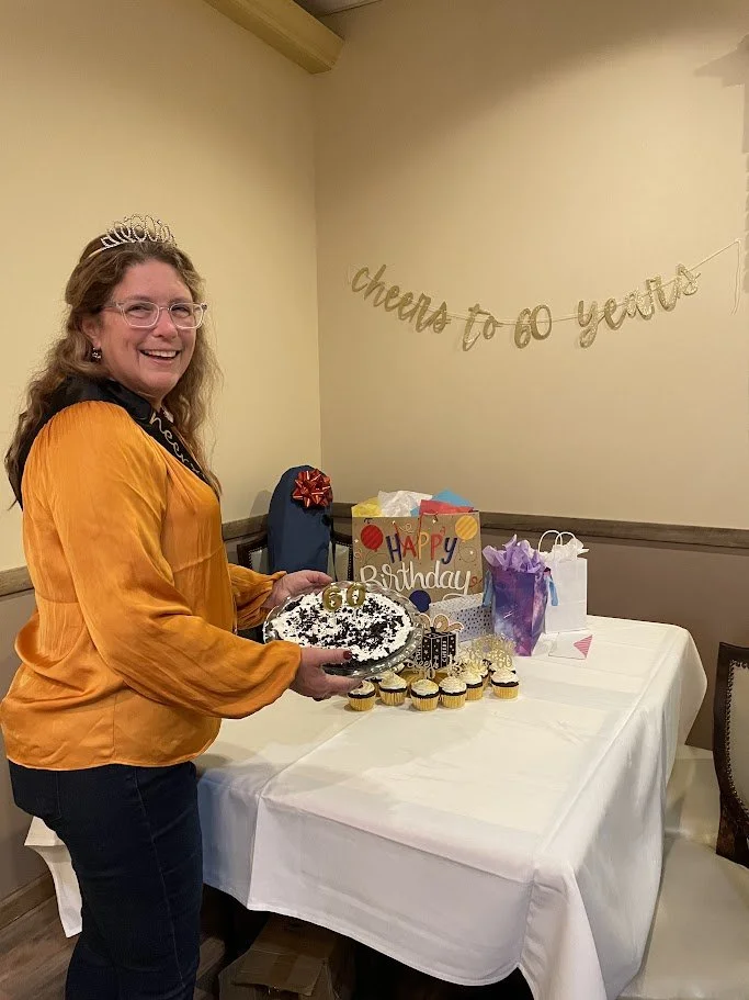 A woman celebrating her birthday with a cake, cupcakes, and gifts on a decorated table, with a 'cheers to go' banner on the wall.