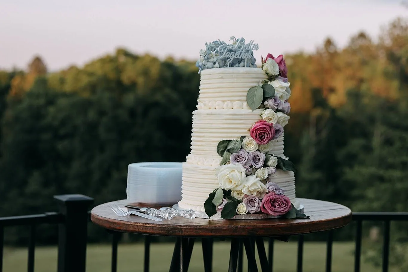 Three-tier wedding cake decorated with pink, white, and purple roses and green leaves, placed on a round wooden table outdoors with a blurred forest background.