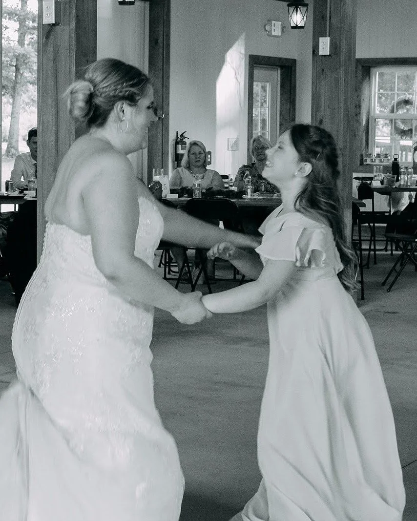 A bride and a young girl are dancing together at a wedding reception in a rustic indoor venue, holding hands and smiling.