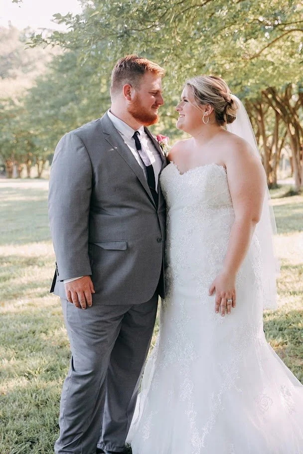 A bride and groom standing close together outdoors during their wedding, looking at each other and smiling. The groom is wearing a gray suit with a black tie, and the bride is in a white strapless wedding gown with a veil. There are trees in the background with sunlight filtering through the leaves.