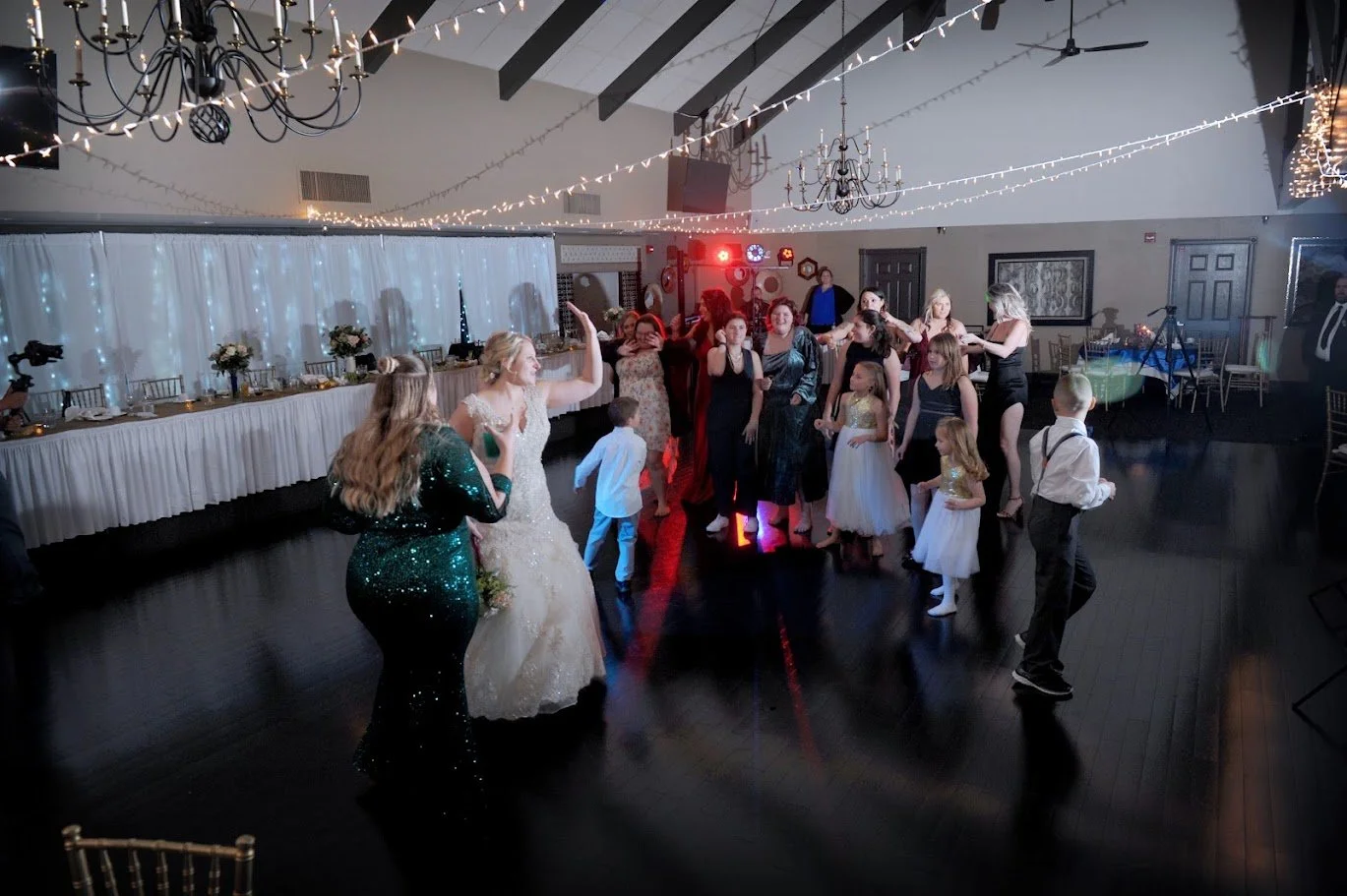 People dancing at a wedding reception in a decorated banquet hall with string lights and chandeliers.
