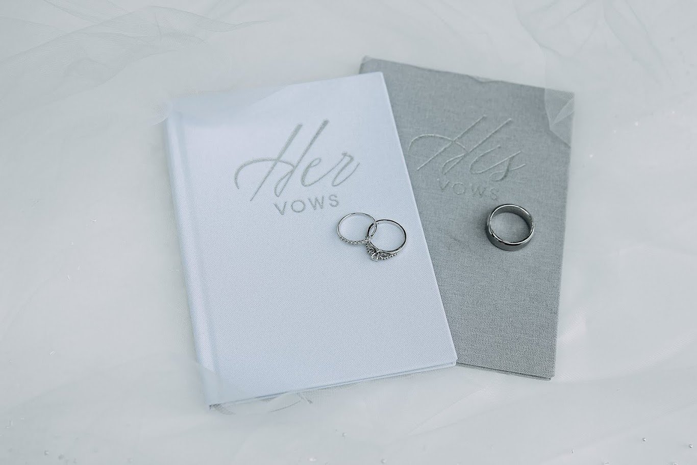 Two wedding vow books, one white and one gray, with rings placed on top. The white book has the words 'Her Vows' in cursive, and the gray book has the words 'His Vows' in cursive.